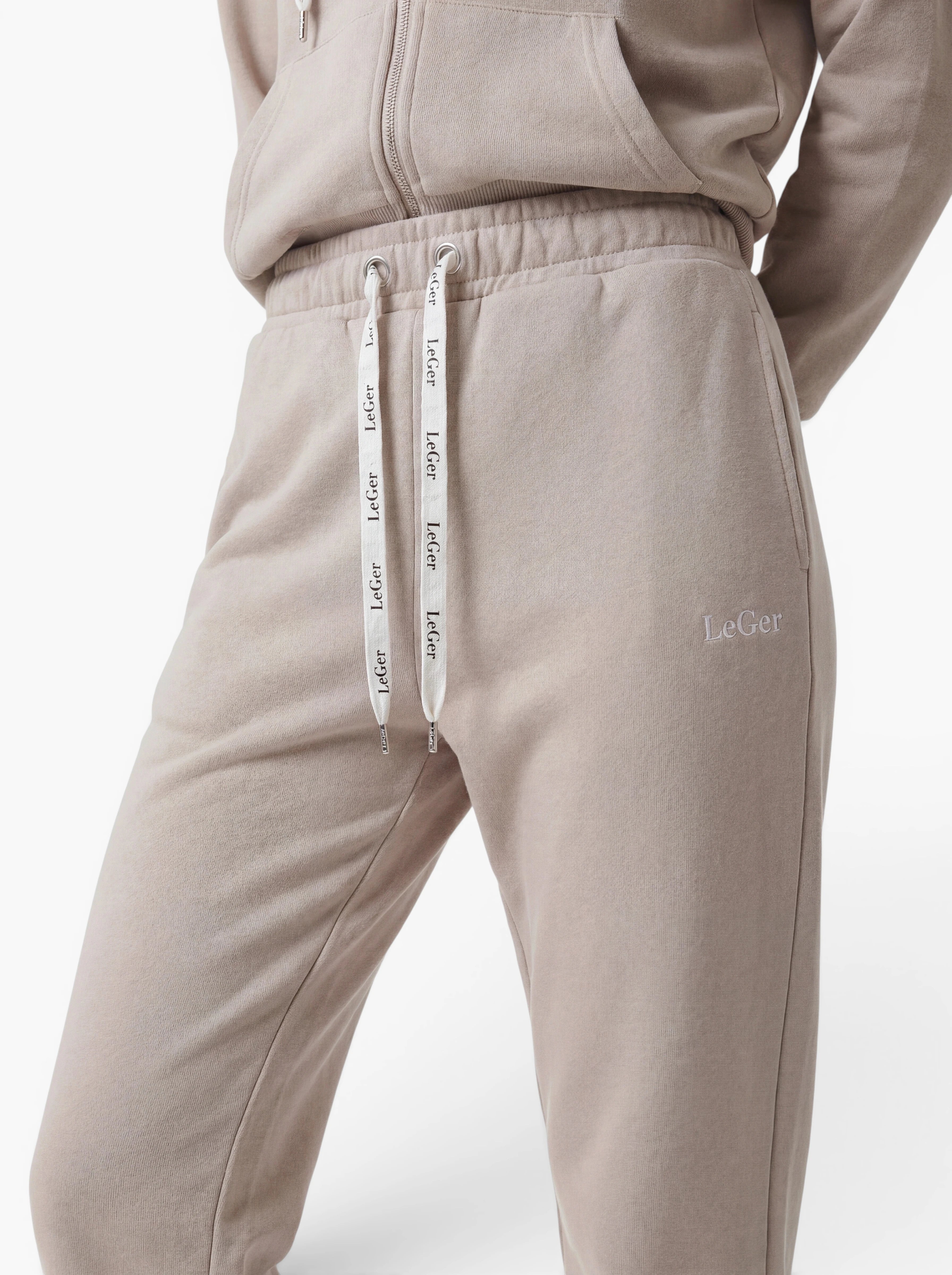 Eine Person trägt die vielseitige Hose „Nela“ von LeGer by Lena Gercke in Beige, kombiniert mit einem passenden Kapuzenpullover. Das „LeGer“-Logo ist sowohl auf der Hose als auch an den Kordeln zu sehen, und die moderne Silhouette sorgt dafür, dass dieses Outfit vor einem weißen Hintergrund stilvoll und vielseitig wirkt.
