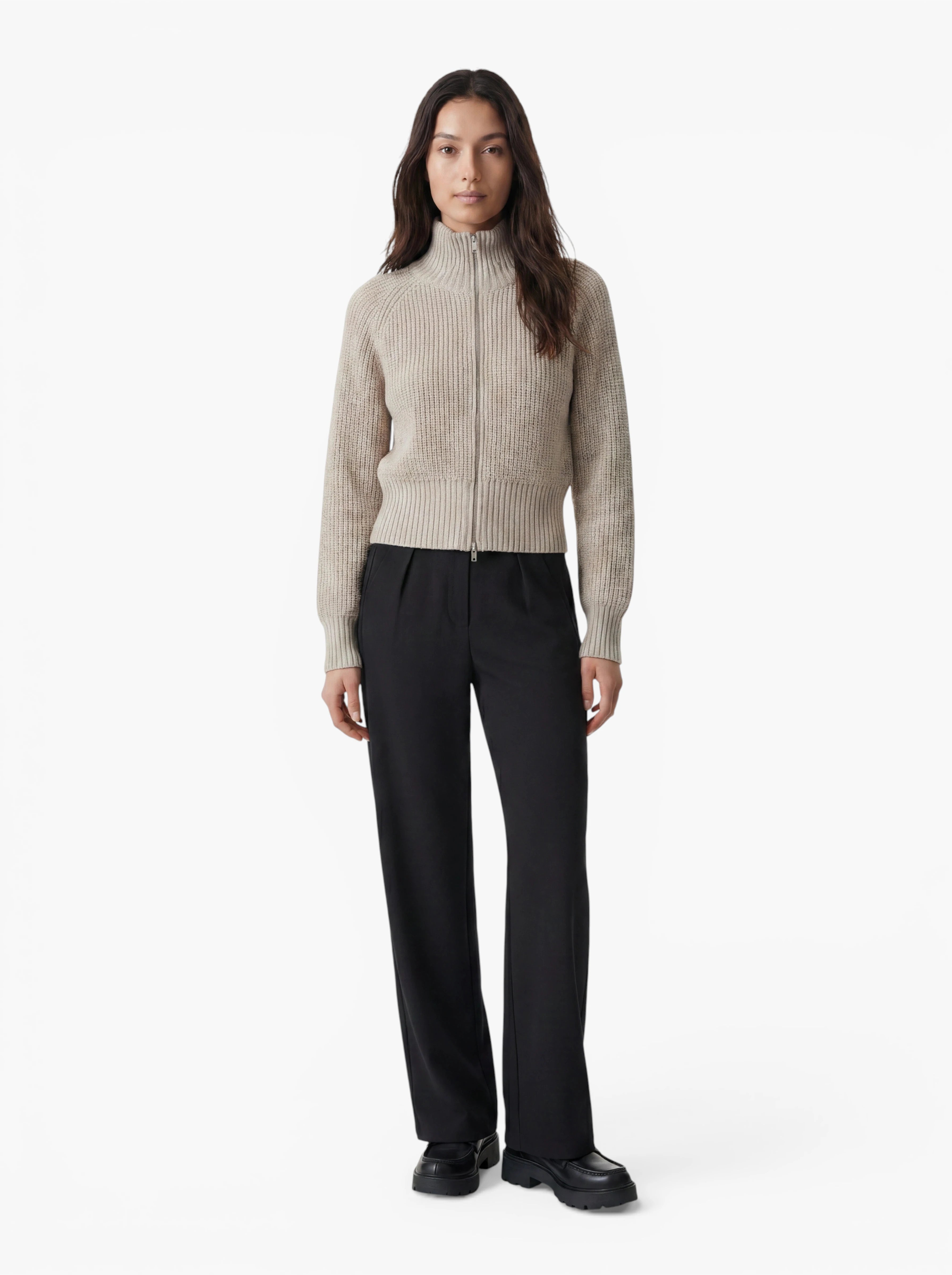 Eine Person steht mit dem Gesicht nach vorne vor einem schlichten weißen Hintergrund und trägt den beigen „Nala“-Pullover von LeGer by Lena Gercke – einen gerippten Pullover mit Reißverschluss –, dazu eine schwarze Hose und schwarze Schuhe.