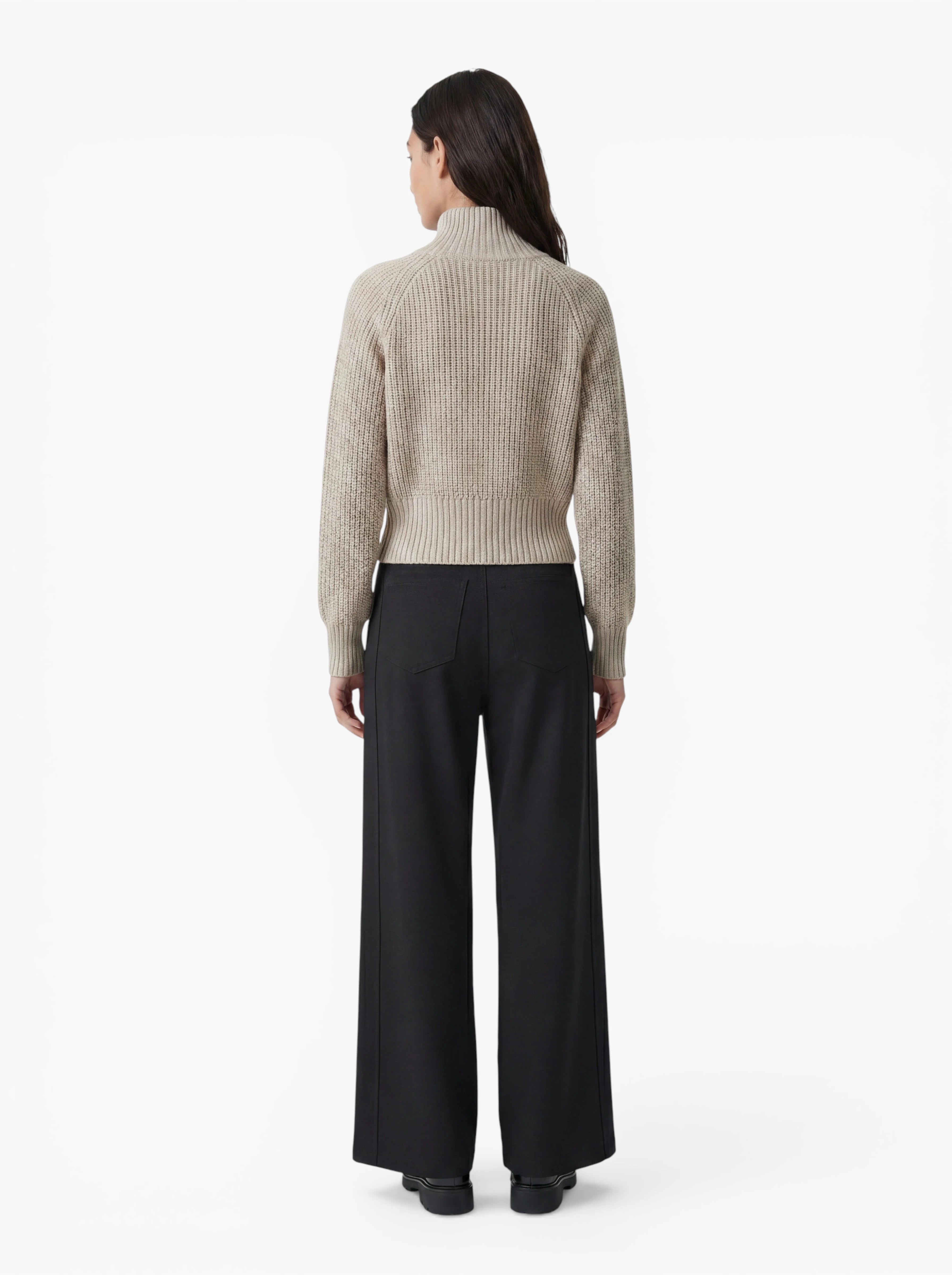 Vor einem schlichten weißen Hintergrund steht eine Person mit langen, dunklen Haaren mit dem Rücken zur Kamera. Sie trägt den Pullover „Nala“ aus beigem Rippstrick von LeGer by Lena Gercke sowie eine schwarze Hose mit weitem Bein.