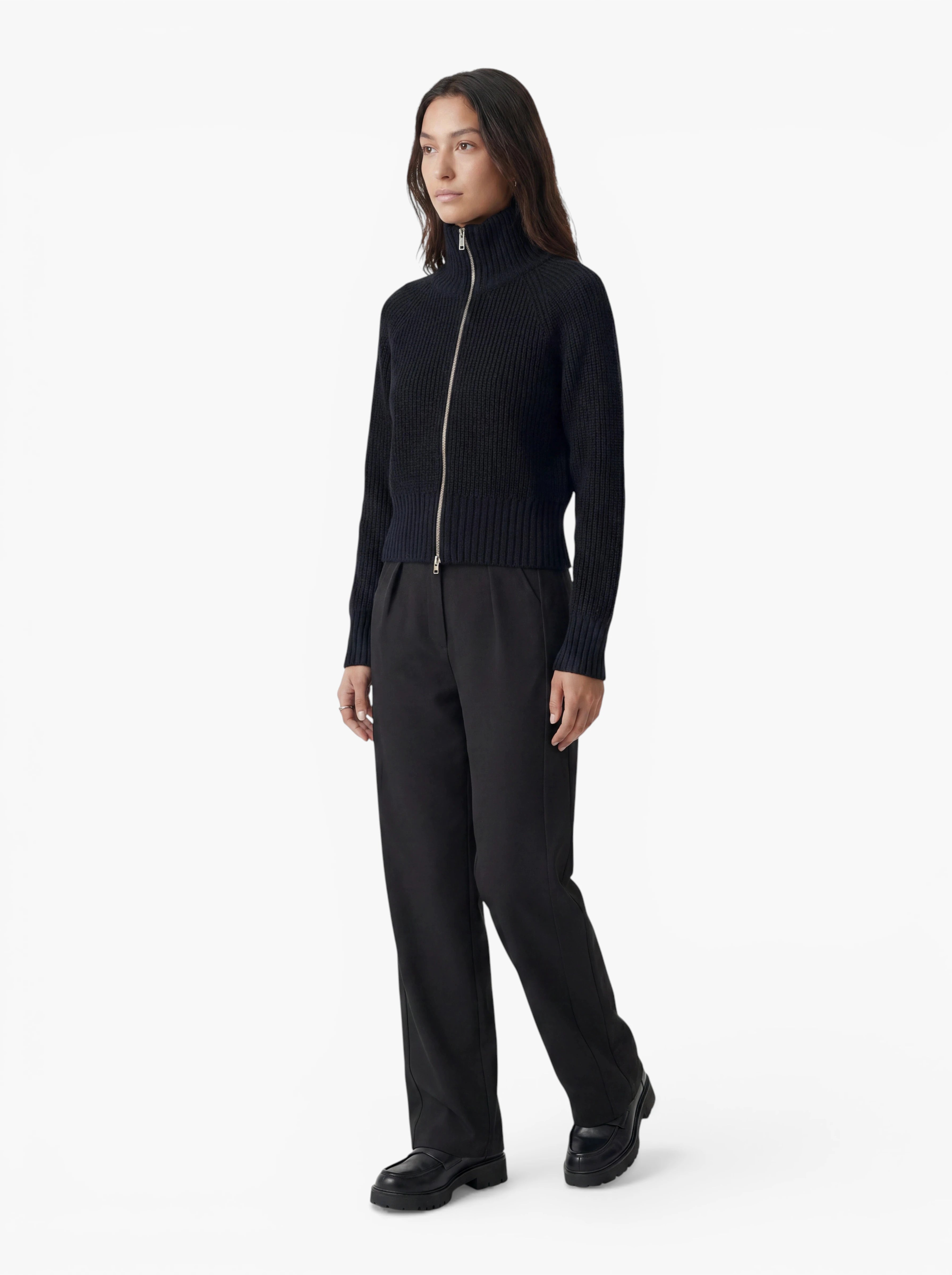 Vor einem schlichten weißen Hintergrund steht eine Person, die den Pullover „Nala“ von LeGer by Lena Gercke trägt – einen schwarzen, gerippten Pullover mit Reißverschluss und entspannter Passform –, dazu eine schwarze Hose mit geradem Bein und schwarze Schuhe.