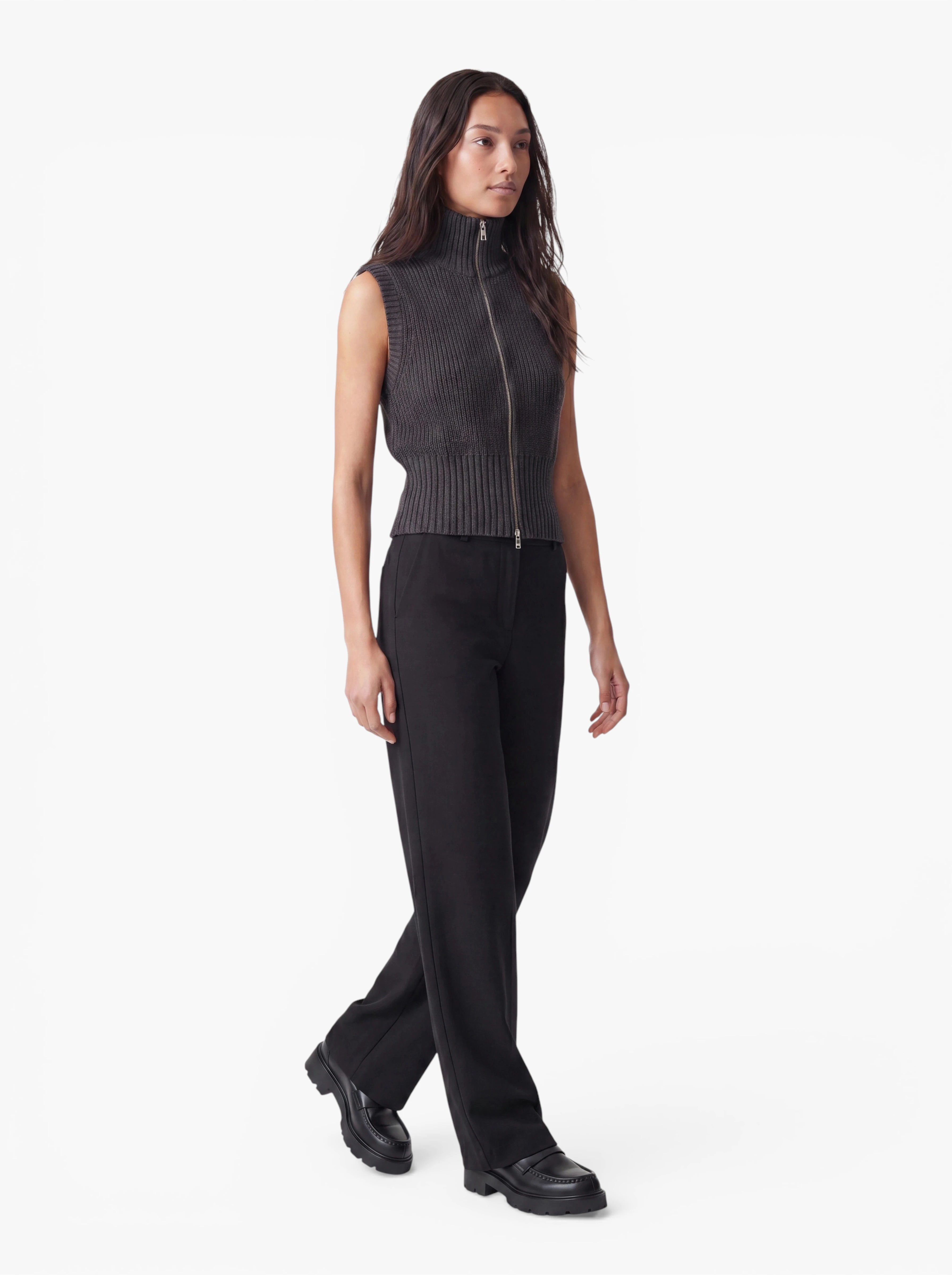 Eine Person mit langen, dunklen Haaren trägt die Weste „Betsy“ von LeGer by Lena Gercke, ein ärmelloses, geripptes Oberteil mit Reißverschluss und minimalistischem Design, dazu eine schwarze Hose und schwarze Schuhe, und läuft vor einem schlichten weißen Hintergrund.