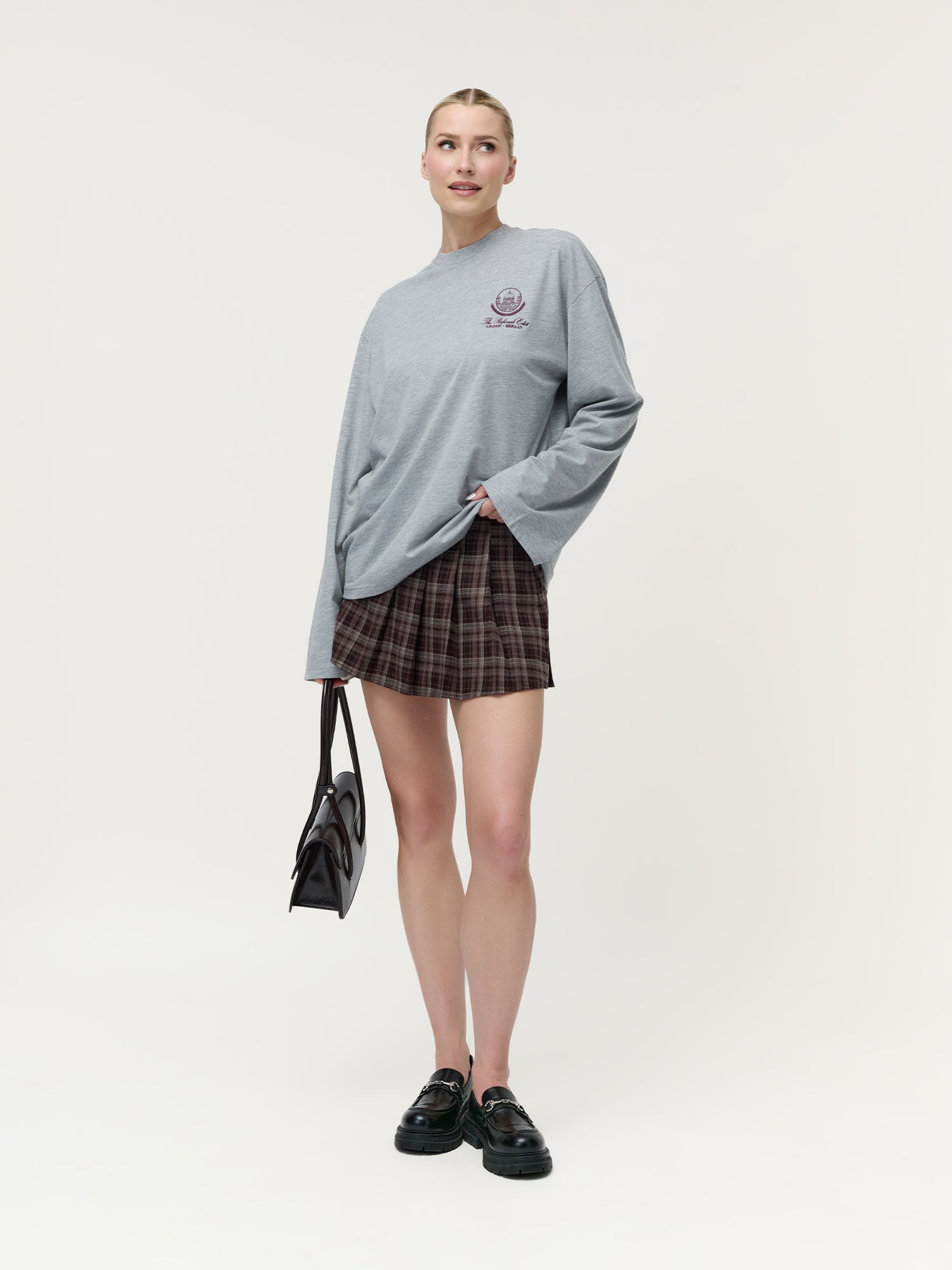 Vor einem weißen Hintergrund steht eine Frau, die das lockere, graue „Briska“-Langarmshirt von LeGer by Lena Gercke trägt, das aus einem weichen Materialmix gefertigt ist. Dazu kombiniert sie einen kurzen Karorock, schwarze Loafer und eine schwarze Handtasche – perfekt für jede Alltagsgarderobe.