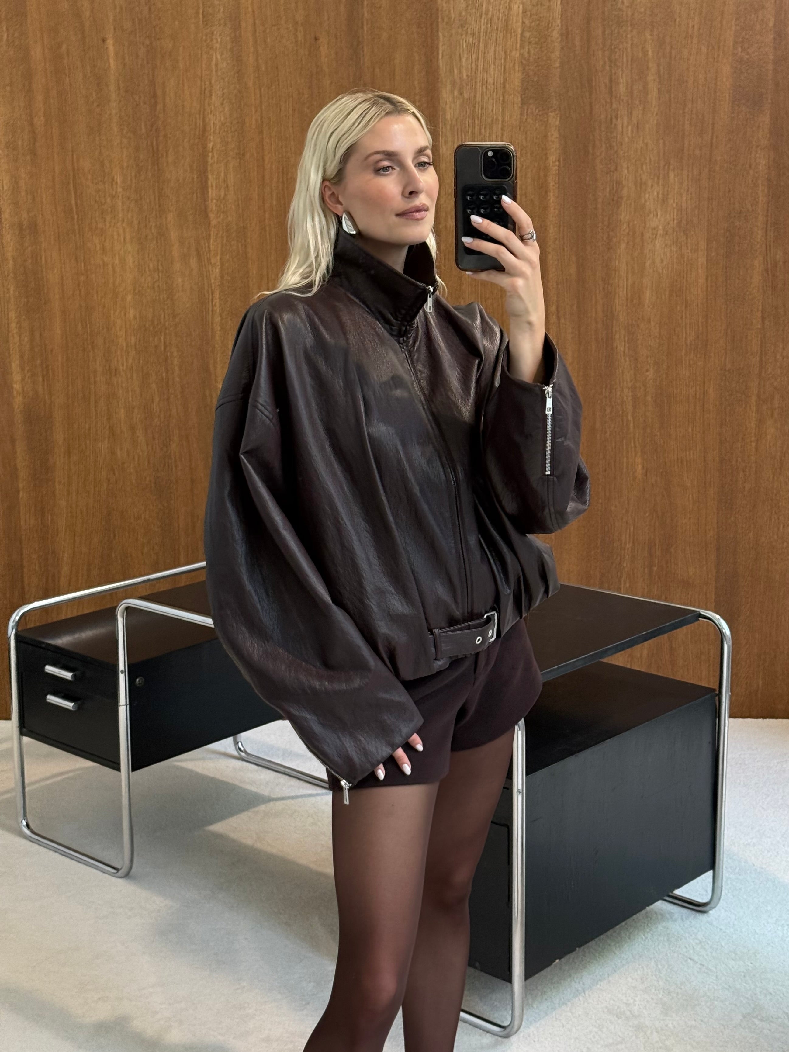 Eine Person mit langen blonden Haaren macht ein Spiegel-Selfie in einer übergroßen „Constance“-Jacke von LeGer by Lena Gercke, die sich durch eine moderne Passform auszeichnet, dazu trägt sie schwarze Shorts und transparente Strumpfhosen und steht vor einem modernen Schreibtisch vor dem Hintergrund einer Holzwand.