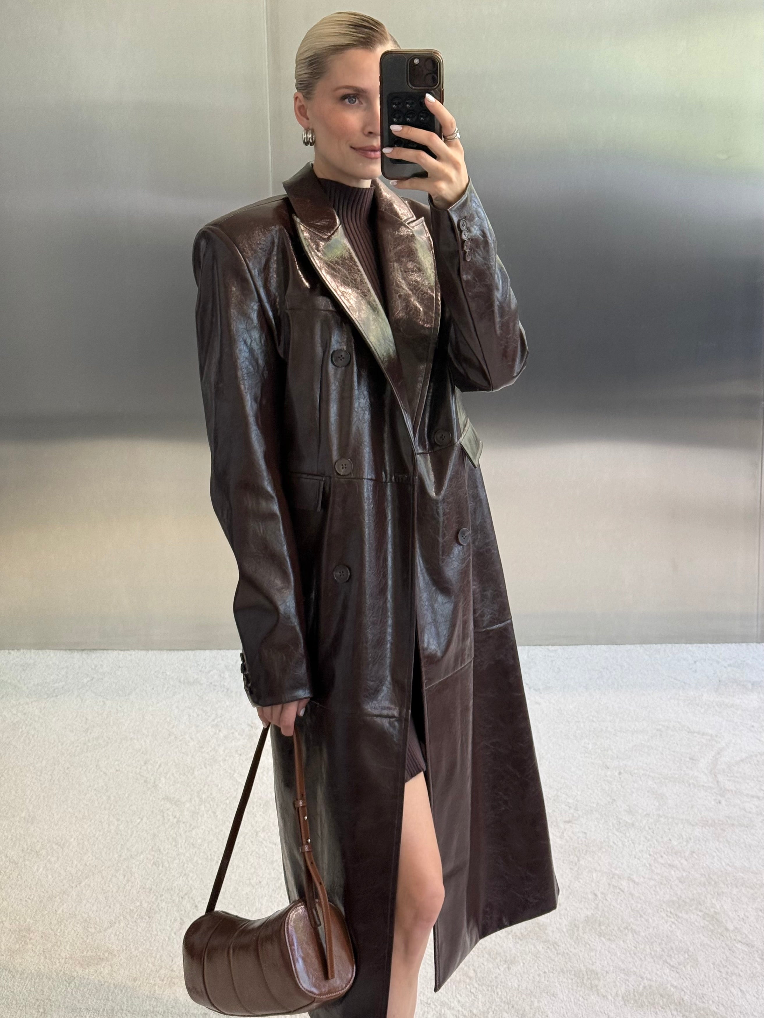 Eine Frau macht ein Spiegel-Selfie, während sie den Mantel „Annika“ von LeGer by Lena Gercke trägt – einen eleganten, dunkelbraunen Damenmantel aus Leder in taillierter Passform –, den sie über einem gerippten Rollkragenkleid trägt, und dabei eine dazu passende braune Handtasche in der Hand hält. Der minimalistische, metallische Hintergrund unterstreicht den raffinierten Look.