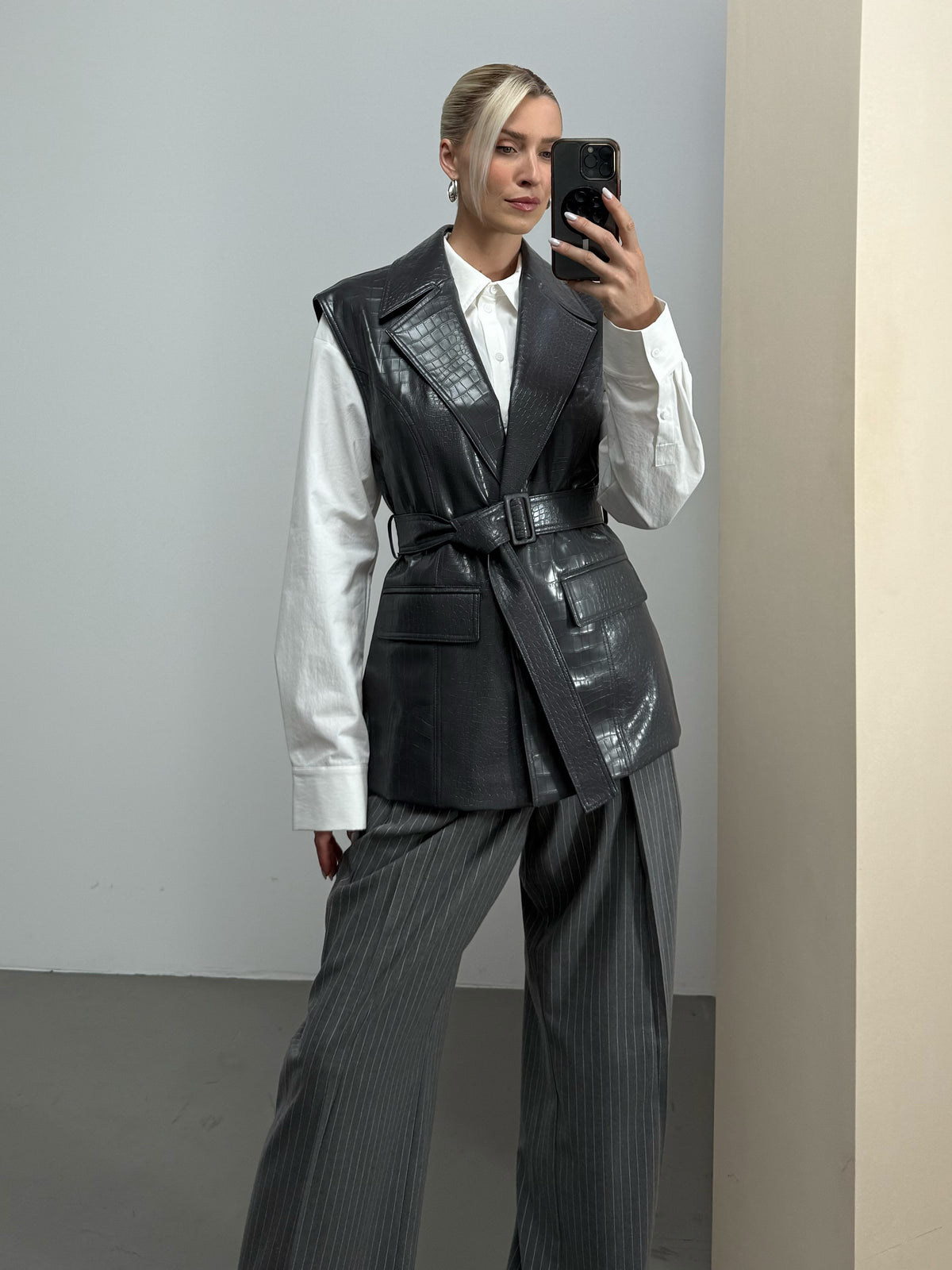 Eine Frau, die ein Spiegel-Selfie macht, trägt ein weißes Hemd, die Weste „Hanne“ von LeGer by Lena Gercke mit einem schwarzen Gürtel sowie eine graue, weit geschnittene Nadelstreifenhose. Sie steht in einem minimalistisch eingerichteten Raum mit neutralen Wänden und einem neutralen Boden.