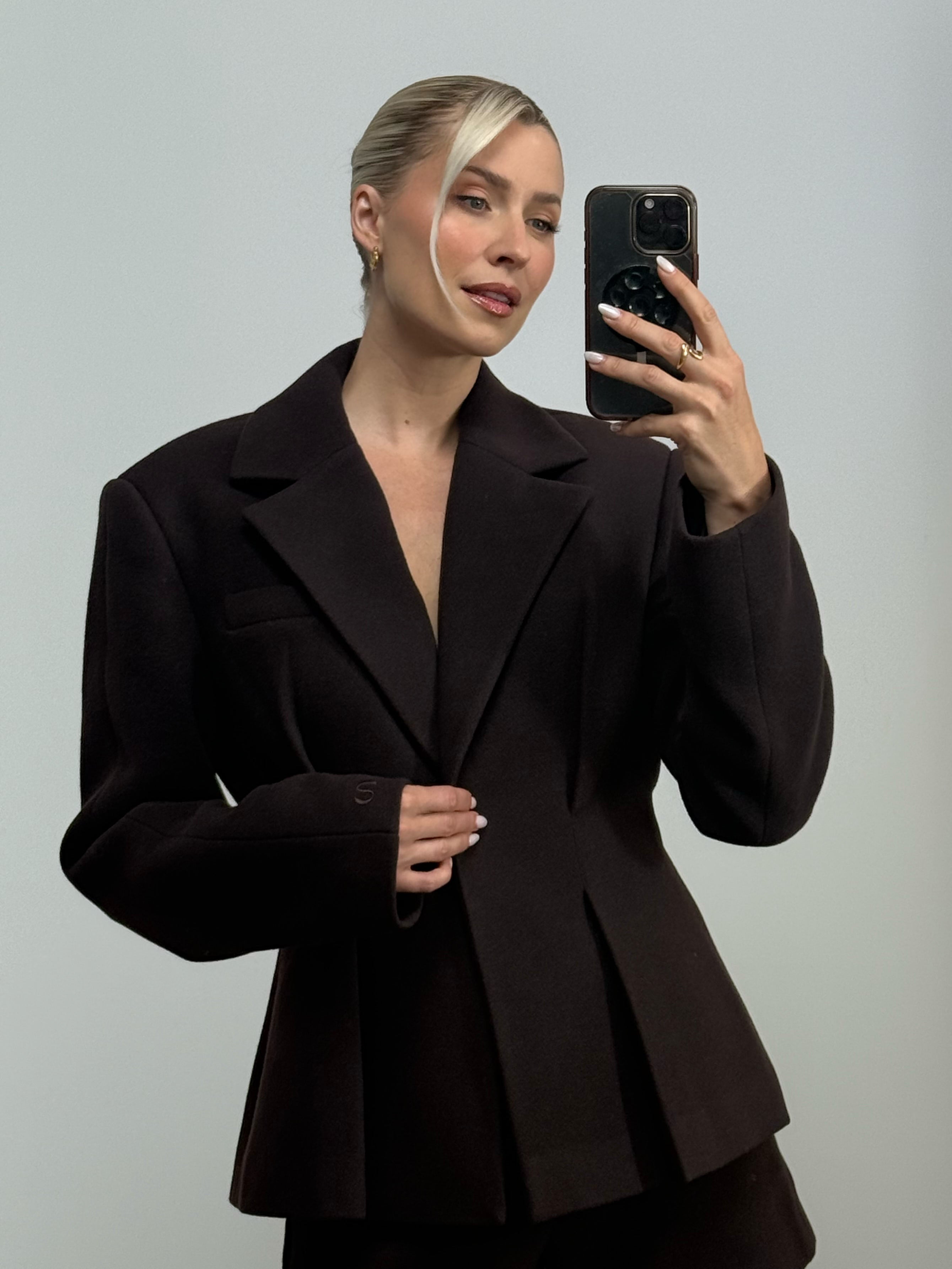 Eine Frau mit blonden Haaren, die zu einem tief sitzenden Dutt gesteckt sind, trägt die Jacke „Jasmin“ von LeGer by Lena Gercke und macht mit ihrem Smartphone vor einem schlichten, hellen Hintergrund ein Spiegel-Selfie.