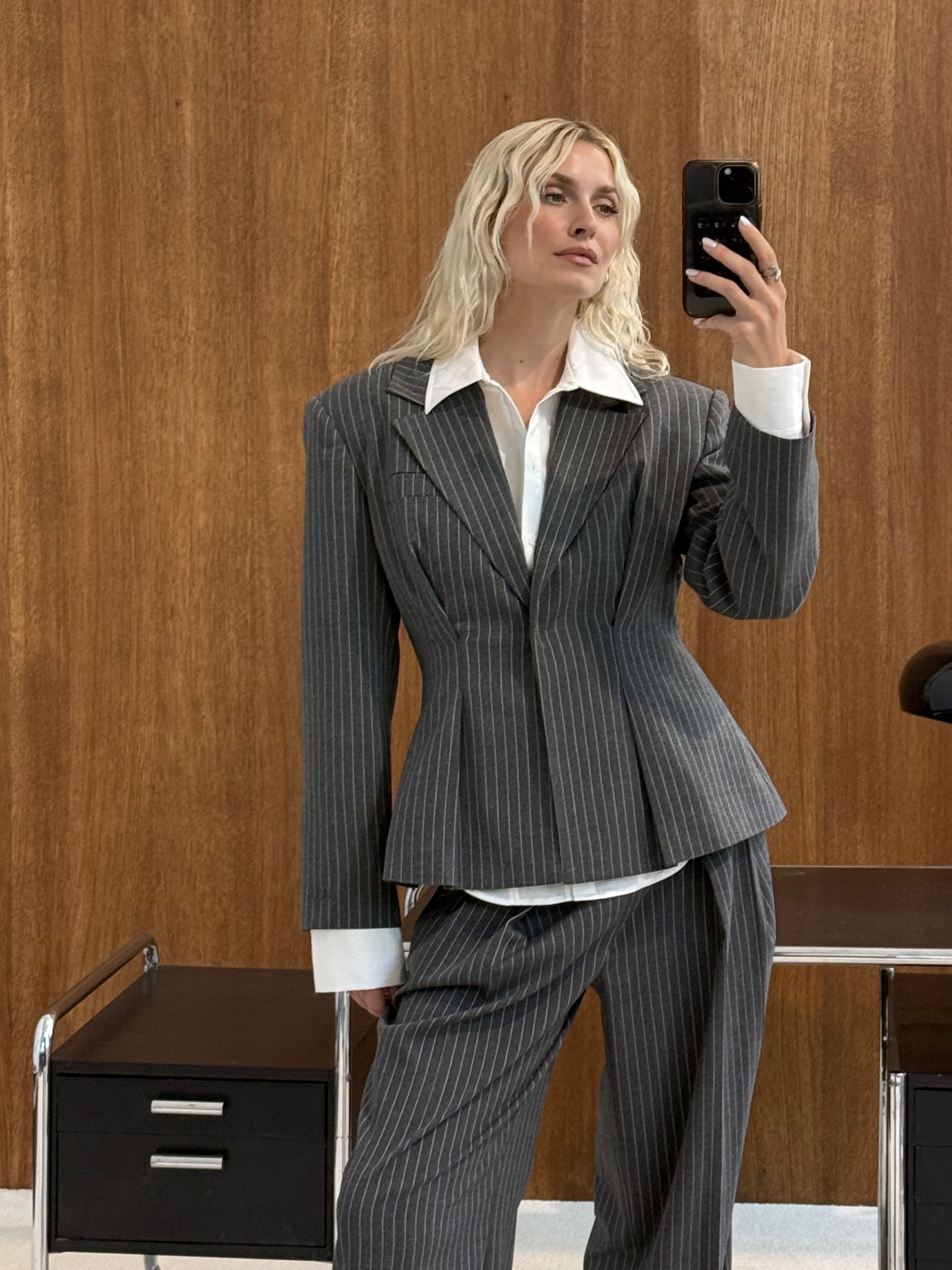 Eine Person, die den Blazer „Sophia“ aus der Kollektion „LeGer by Lena Gercke“ im klassischen Nadelstreifen-Design trägt, macht ein Spiegel-Selfie in einer Büroumgebung, wobei im Hintergrund eine Holzwand, ein schwarzer Stuhl und ein Schubladenschrank zu sehen sind.