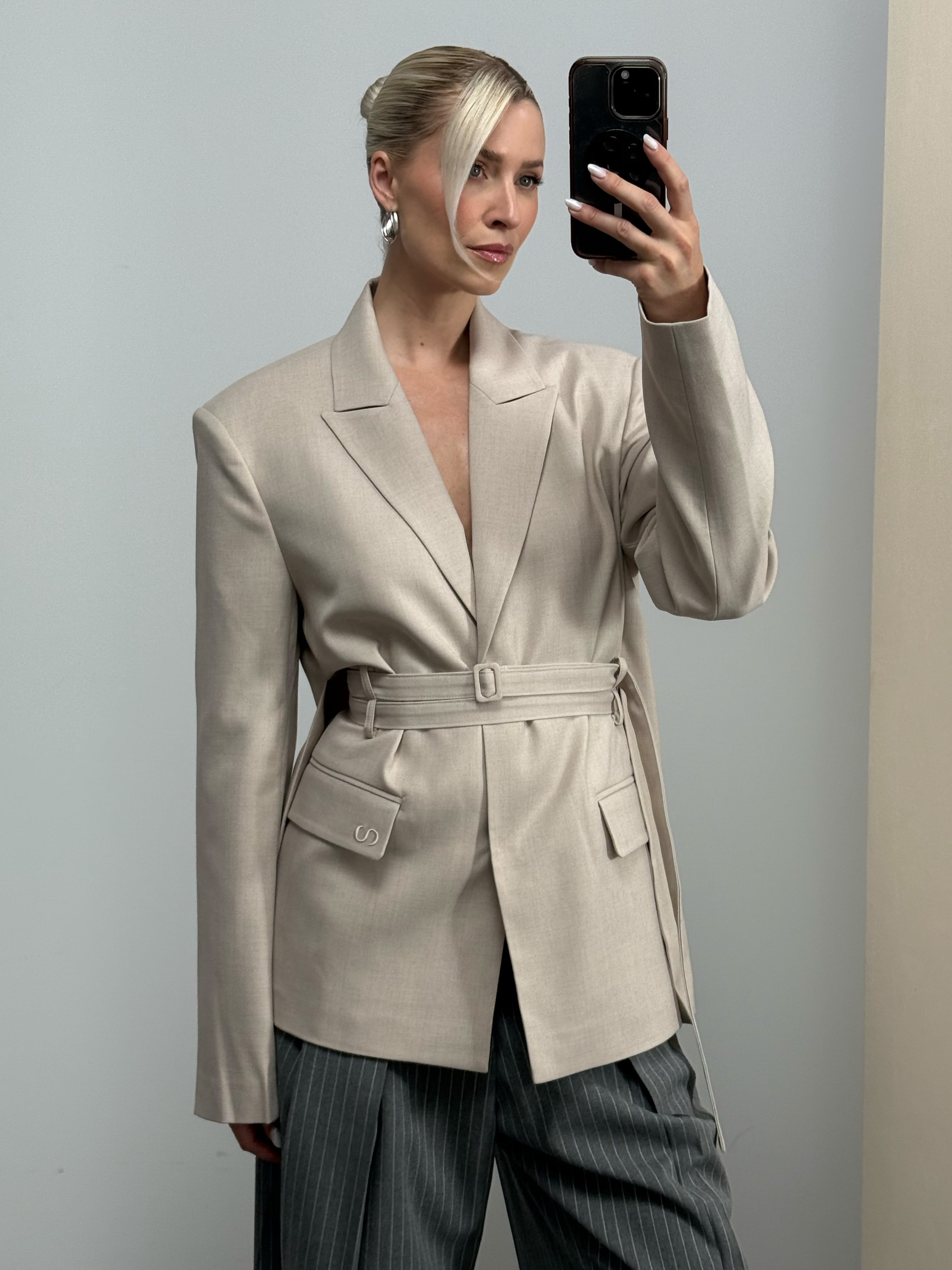 Eine Frau, die den beigen Blazer „Frances“ von LeGer by Lena Gercke trägt – der sich durch einen modernen Schnitt und einen doppelten Taillengürtel auszeichnet –, macht ein Spiegel-Selfie. Ihr Haar ist zu einem Dutt gestylt, der seitlich geflochtene Pony fällt ihr ins Gesicht, und der Hintergrund ist schlicht und neutral.