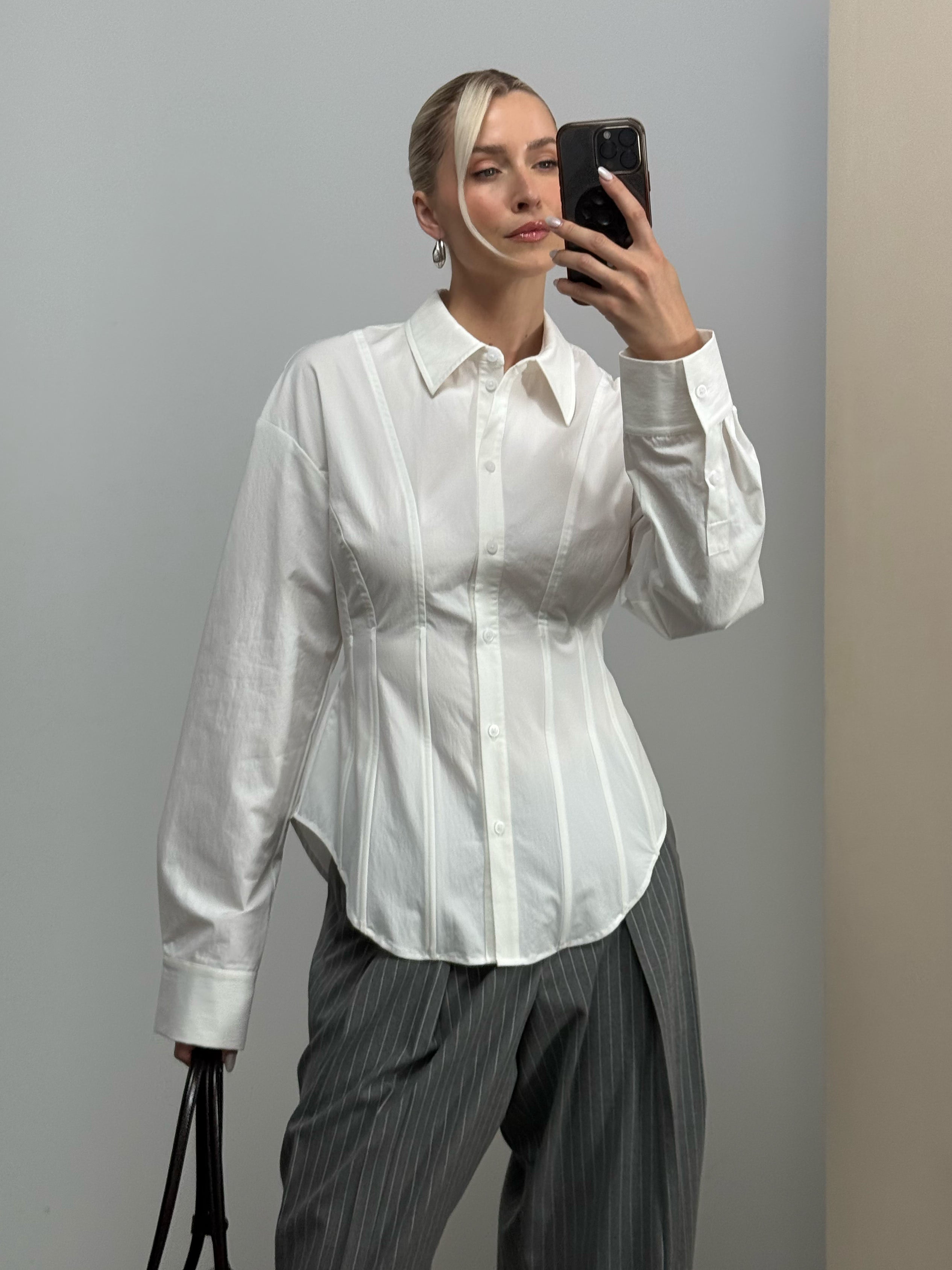 Eine Person trägt die taillierte „Alannah“-Bluse von LeGer by Lena Gercke sowie eine graue Nadelstreifenhose, macht ein Spiegel-Selfie und hält eine schwarze Tasche in der Hand. Der Hintergrund ist schlicht und besteht aus neutralen Wänden.