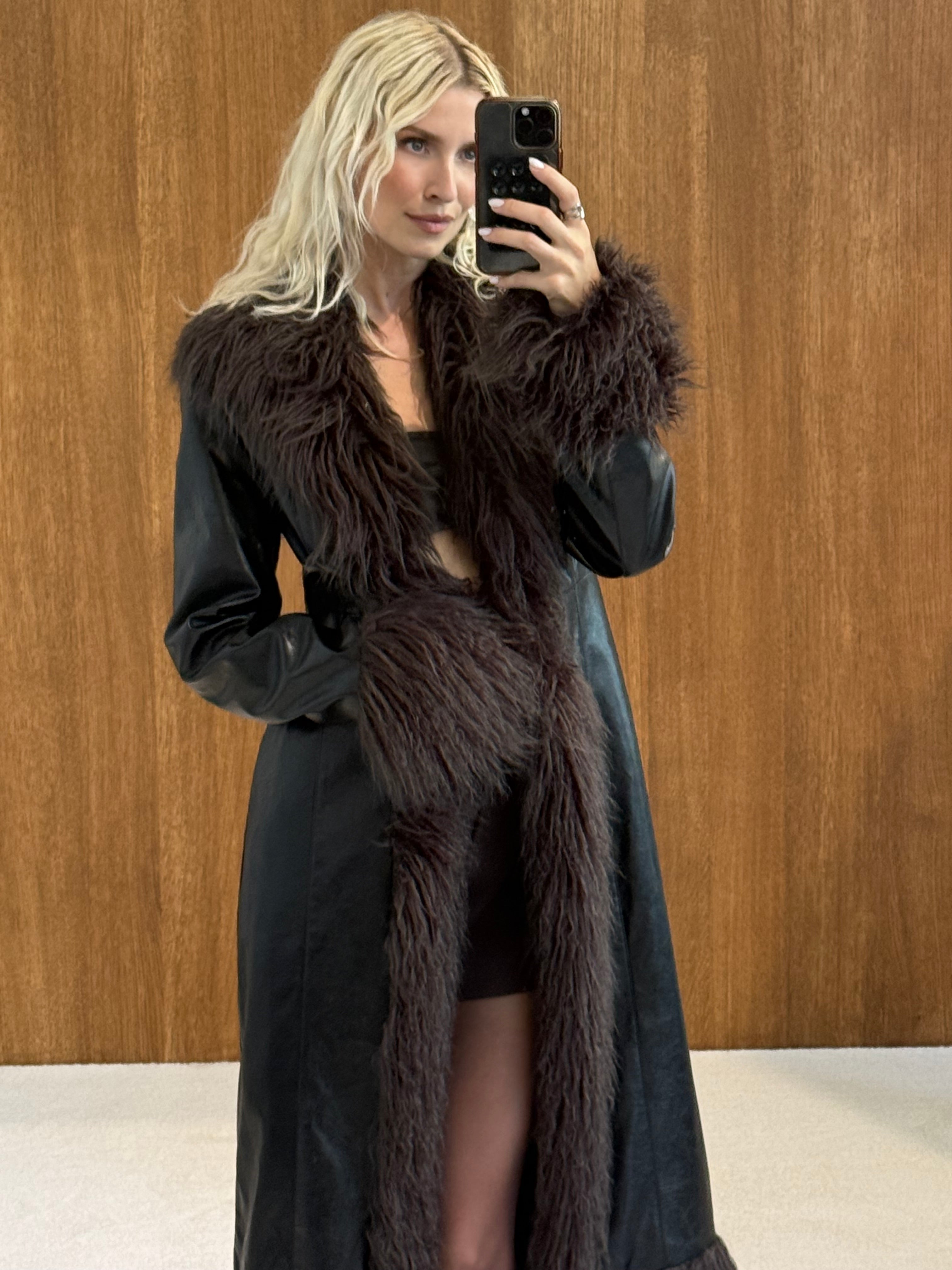 Eine Person mit langen blonden Haaren macht ein Spiegel-Selfie. Sie trägt den taillierten Damenmantel „Teena“ von LeGer by Lena Gercke – einen schwarzen Mantel mit großen, dunkelbraunen Kunstfellbesätzen an Kragen und Manschetten – und steht dabei vor einer Holzwand.