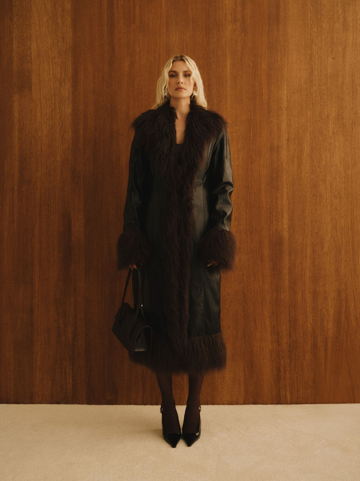 Eine Person steht vor einer Holzwand und trägt den langen, maßgeschneiderten „Teena“-Mantel von LeGer by Lena Gercke mit dunklem Kunstpelzbesatz sowie schwarze Pumps; in der Hand hält sie eine schwarze Handtasche. Der Look und die Pose strahlen eine stilvolle, elegante Atmosphäre aus, die vom legendären „Teena“-Mantel inspiriert ist.