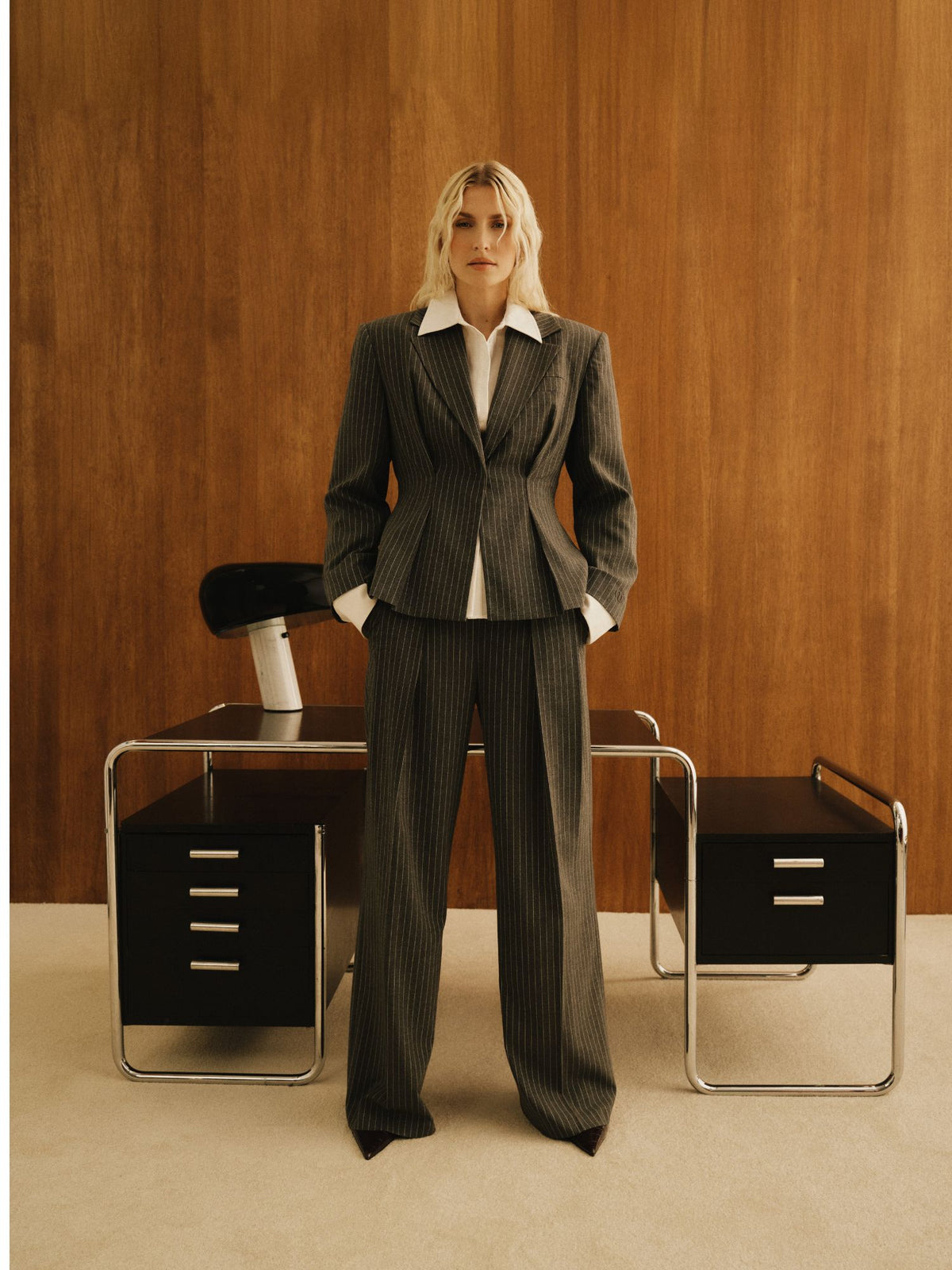 Im Look von LeGer by Lena Gercke: Eine Person trägt die Hose „Sophia“ zu einem Nadelstreifenanzug und steht selbstbewusst – die Hände in den Taschen – vor einem Vintage-Schreibtisch und einer holzvertäfelten Wand, ergänzt durch einen modernen schwarzen Stuhl und eine minimalistische Büroeinrichtung.