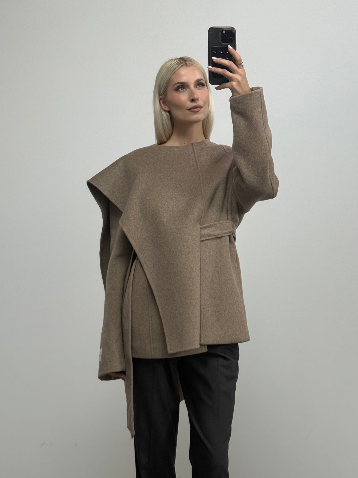 Eine Person mit langen blonden Haaren macht vor einer schlichten weißen Wand ein Spiegel-Selfie. Sie trägt dabei die Jacke „Elva“ von LeGer by Lena Gercke – ein asymmetrisches, braunes Modell im Cape-Stil – sowie eine schwarze Hose.