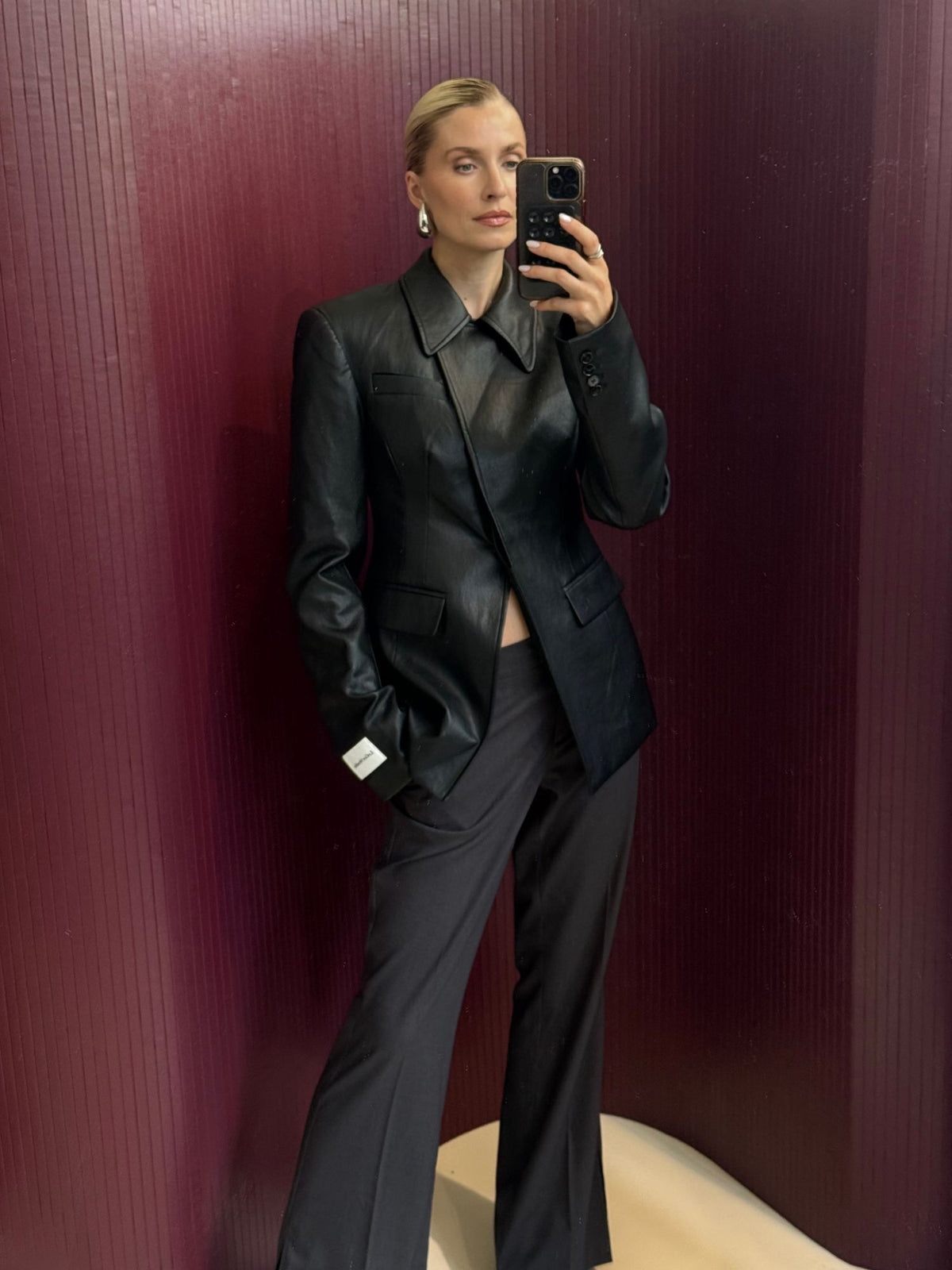 Eine Person, die den Blazer „Orelia“ von LeGer by Lena Gercke in schwarzem Leder-Look mit taillierter, schmaler Passform trägt, kombiniert mit einer dunklen Hose und Ohrringen, macht ein Spiegel-Selfie vor einem strukturierten, weinroten Hintergrund.