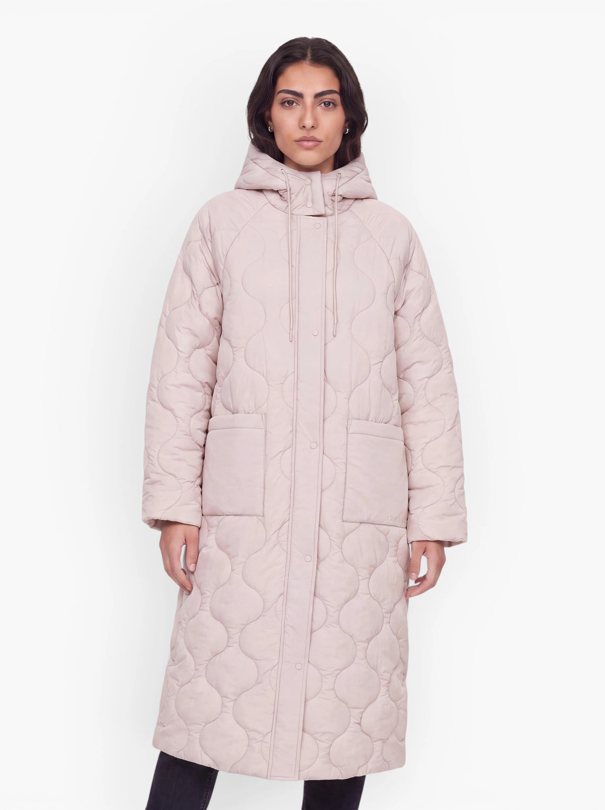 Eine Frau, die den langen, hellrosa, gesteppten Mantel „Fray“ (langer Steppmantel) von LeGer by Lena Gercke trägt – der sich durch einen Oversize-Schnitt, eine Kapuze und große Vordertaschen auszeichnet –, steht vor einem schlichten weißen Hintergrund.