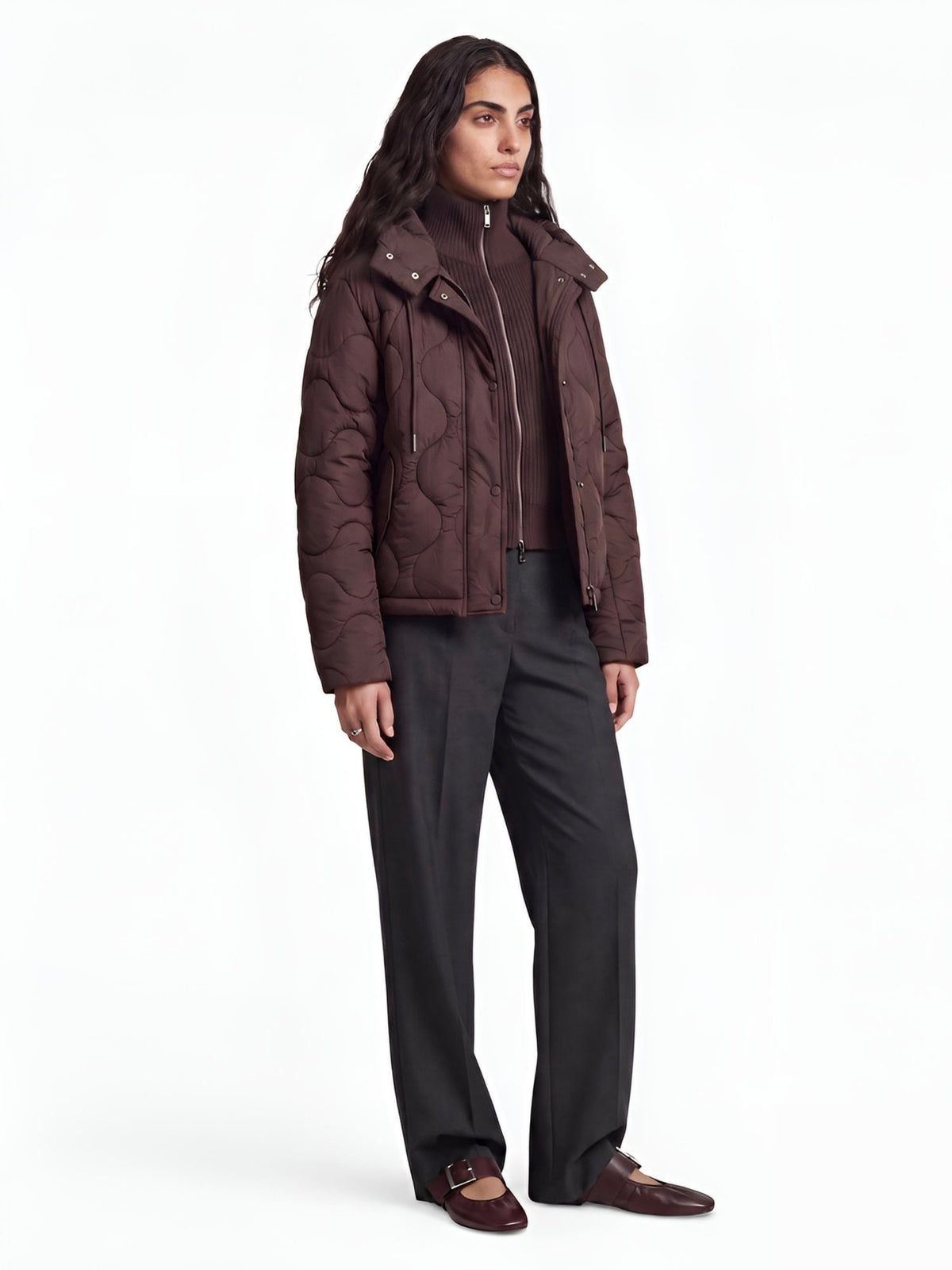 Eine Person mit langen, dunklen Haaren trägt die braune Steppjacke „Fray“ von LeGer by Lena Gercke im Oversize-Schnitt, einen braunen Rollkragenpullover, eine weite, dunkle Hose und braune Schuhe und steht vor einem schlichten weißen Hintergrund.