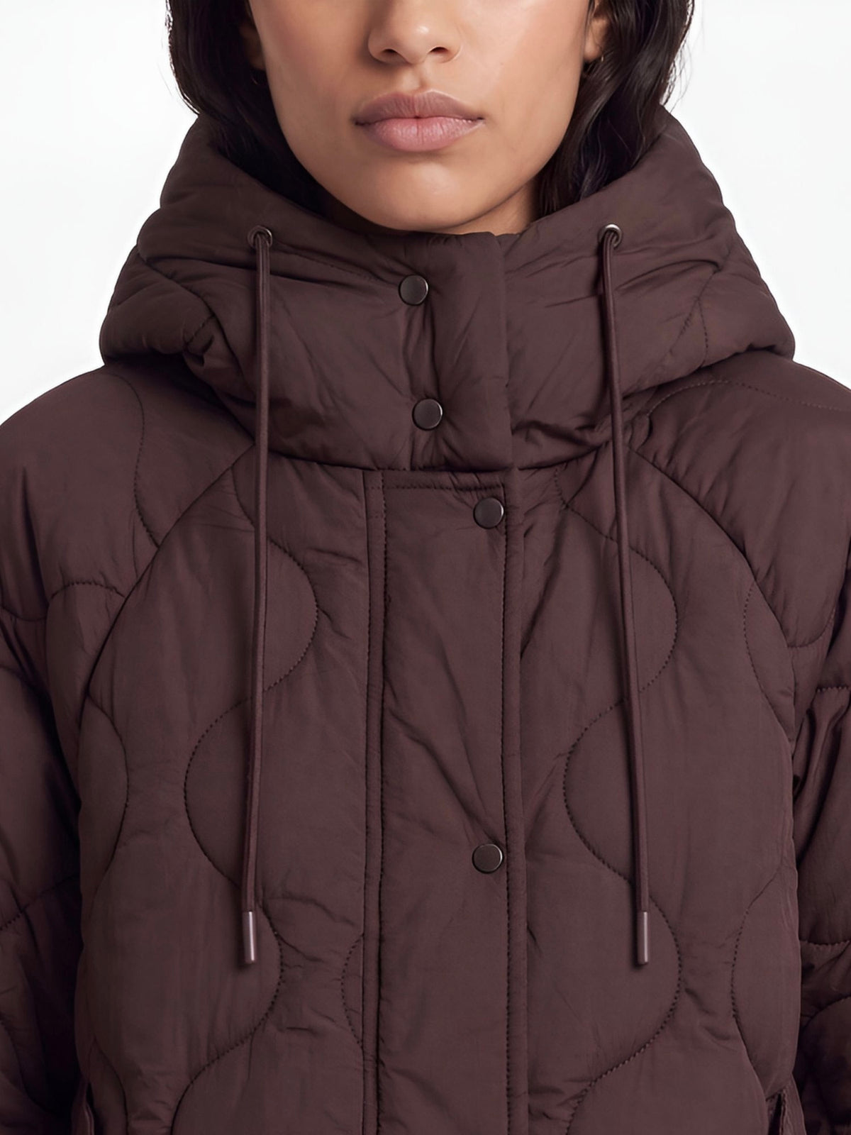 Eine Person, die die Jacke „Fray“ von LeGer by Lena Gercke trägt – eine braune, gesteppte Oversize-Jacke mit hohem Kragen und Kapuze, die mit Knöpfen und Kordelzügen versehen ist. Das Bild zeigt den Oberkörper und einen Teil des Gesichts vor einem neutralen Hintergrund.