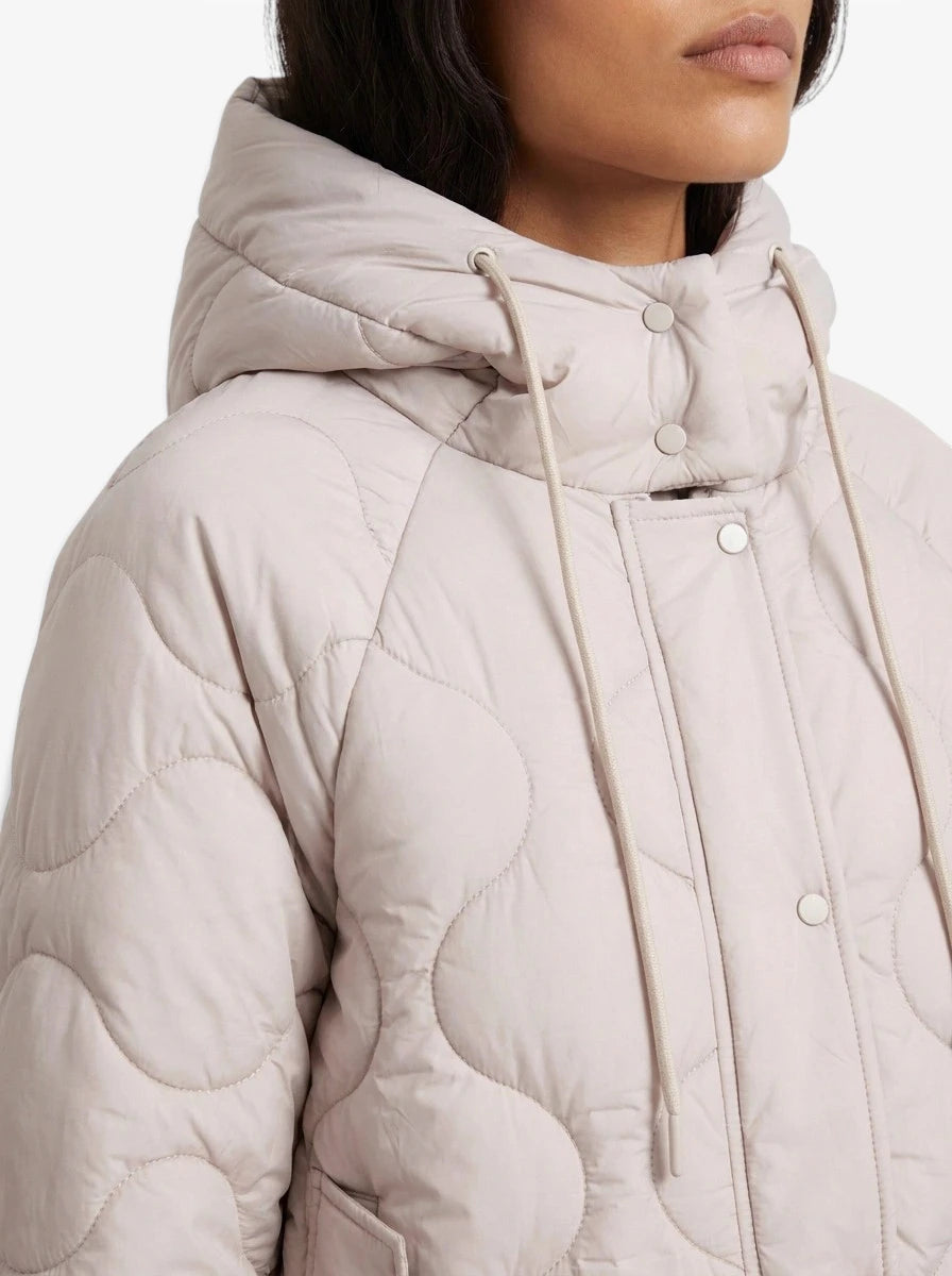 Eine Person ist zu sehen, die die Jacke „Fray“ von LeGer by Lena Gercke trägt – eine hellbeige, oversized Steppjacke mit Kapuze, Druckknopfverschluss und Kordelzug. Nur der untere Teil ihres Gesichts und der obere Teil ihres Oberkörpers sind zu sehen.