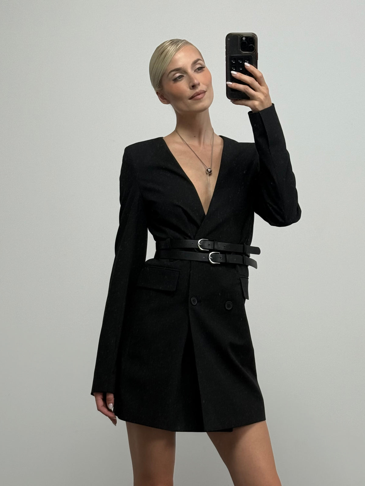 Eine Person steht vor einem schlichten Hintergrund und trägt das stilvolle Kleid „Julie“ in Anthrazit von LeGer by Lena Gercke mit zweireihiger Knopfleiste und doppelten Gürteln. Sie macht mit ihrem Handy ein Spiegel-Selfie.
