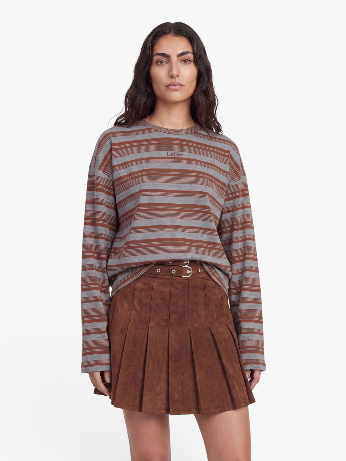 Eine Frau, die das Langarmshirt „Vivian“ von LeGer by Lena Gercke mit grau-braunen Streifen und einen braunen Faltenrock trägt, steht mit dem Gesicht nach vorne vor einem schlichten weißen Hintergrund.