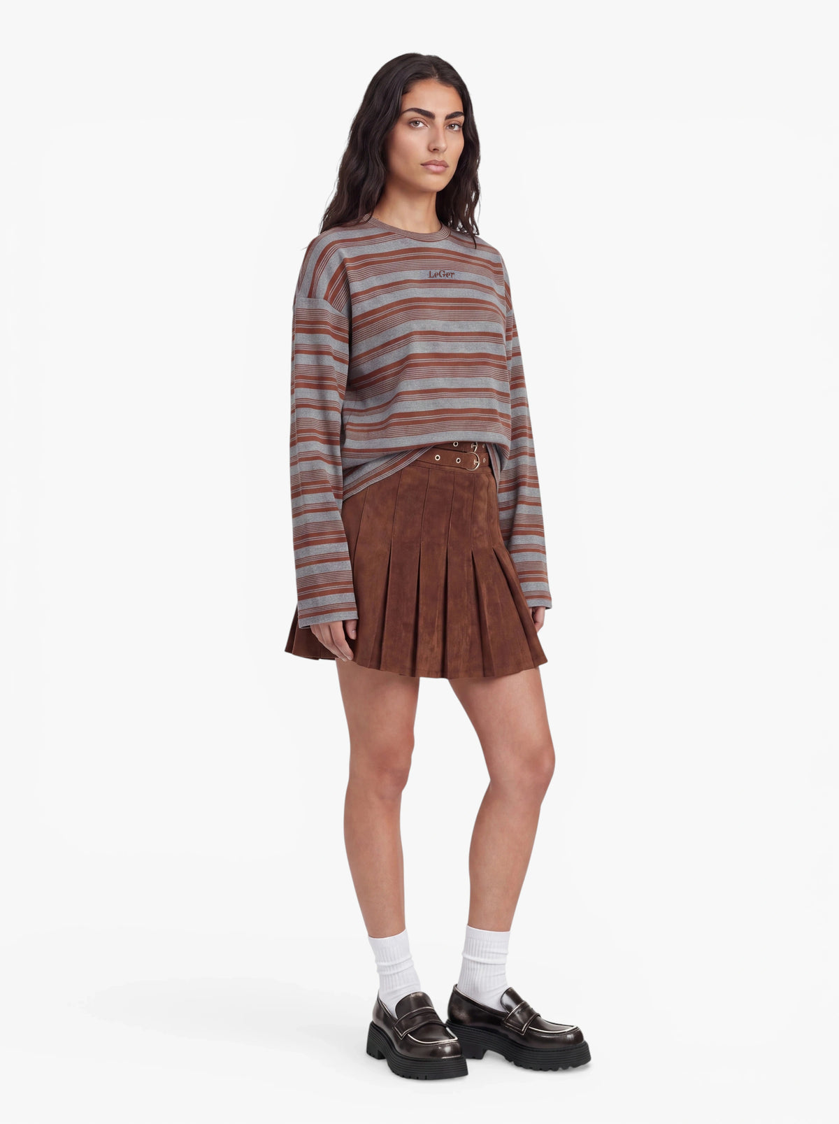 Eine Person steht vor einem schlichten weißen Hintergrund und trägt einen braunen Faltenrock, das Modell „Vivian“ von LeGer by Lena Gercke – ein Langarmshirt mit grau-braunen Streifen, weiße Socken und schwarze Loafer.