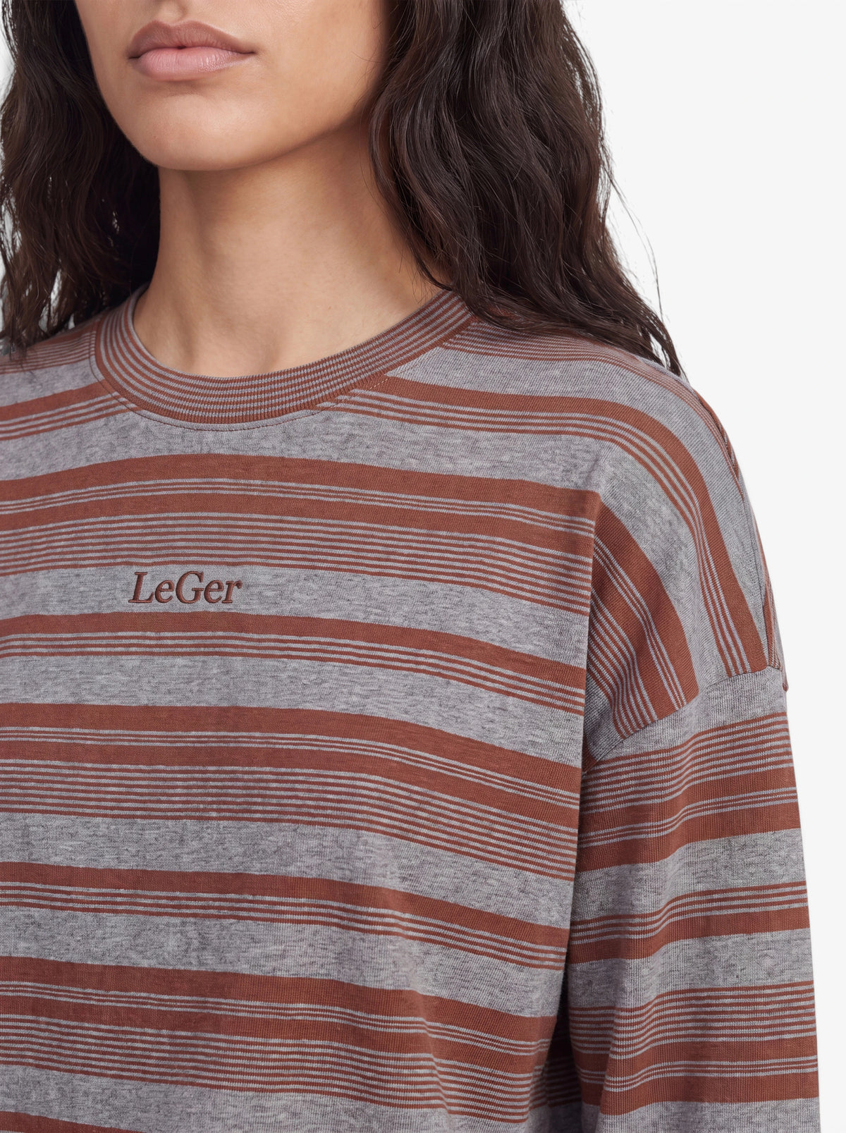 Eine Frau mit langen, welligen, dunklen Haaren präsentiert das Langarmshirt „Vivian“ von LeGer by Lena Gercke, das sich durch grau-braune Streifen und ein gesticktes „LeGer“-Logo auf der Brust auszeichnet. Zu sehen sind nur ihr unterer Gesichtsbereich, ihr Hals und ihr oberer Oberkörper.