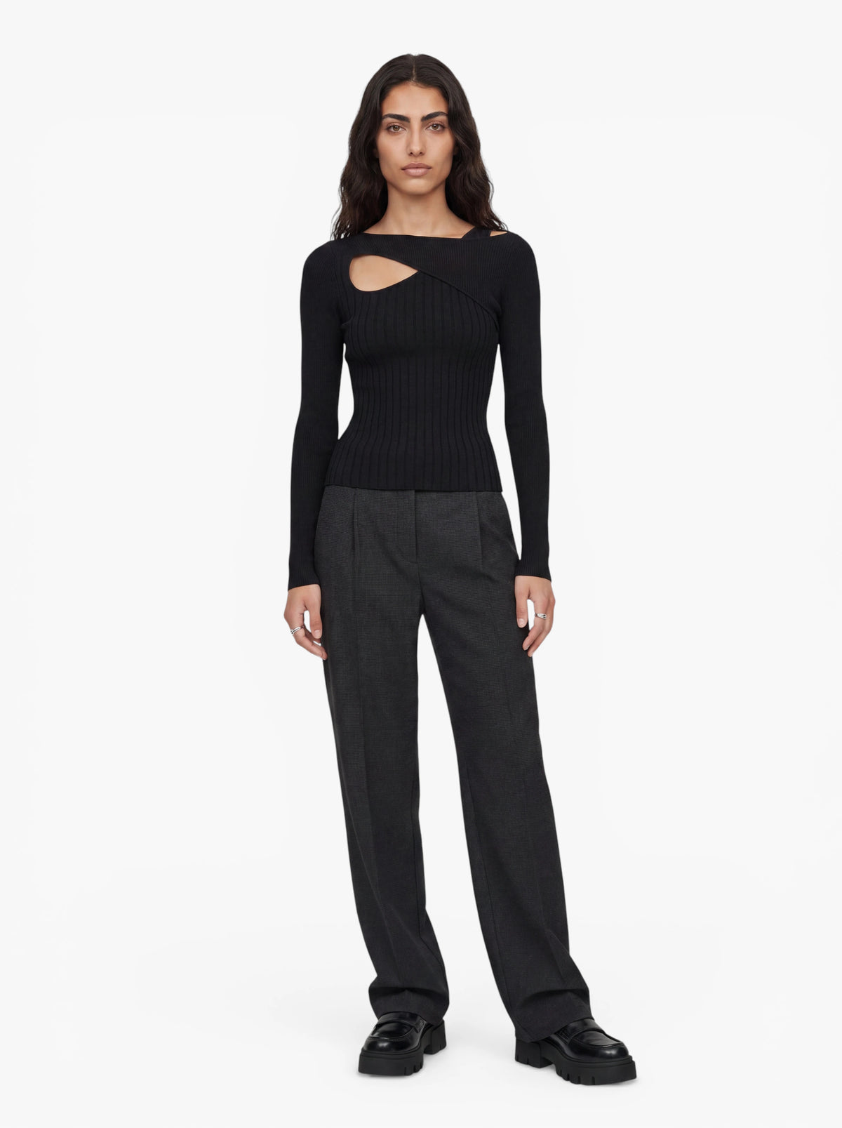 Vor einem schlichten weißen Hintergrund steht eine Frau mit Blick nach vorne und trägt den Pullover „Selina“ von LeGer by Lena Gercke – einen schwarzen, gerippten Strickpullover mit Ausschnitt und asymmetrischem Halsausschnitt –, dazu eine graue Hose mit hoher Taille und schwarze Plateauschuhe.
