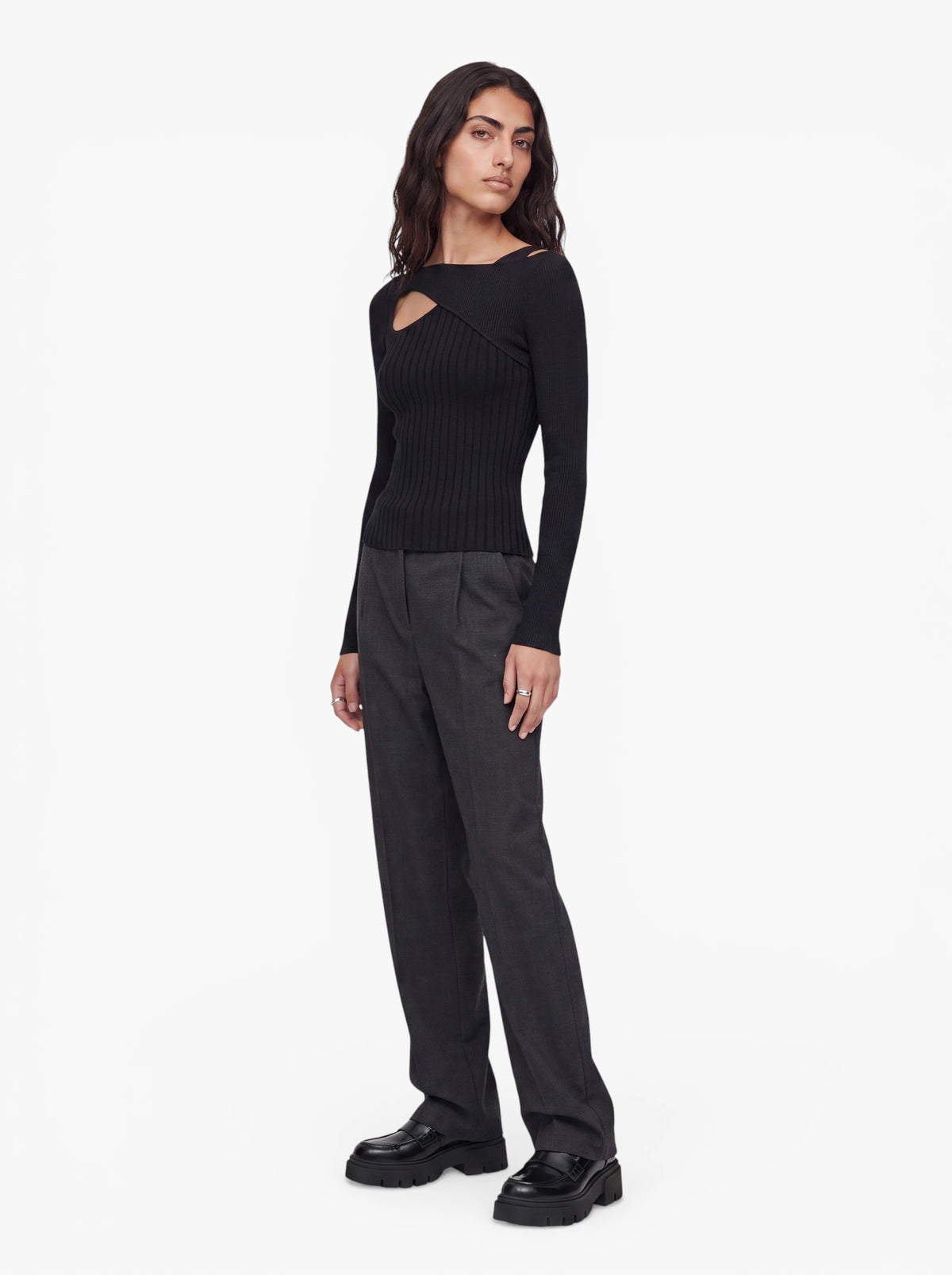 Eine Person mit langen, dunklen Haaren, die den Pullover „Selina“ von LeGer by Lena Gercke trägt, kombiniert mit einer dunkelgrauen Hose und schwarzen Schuhen, steht vor einem schlichten weißen Hintergrund.