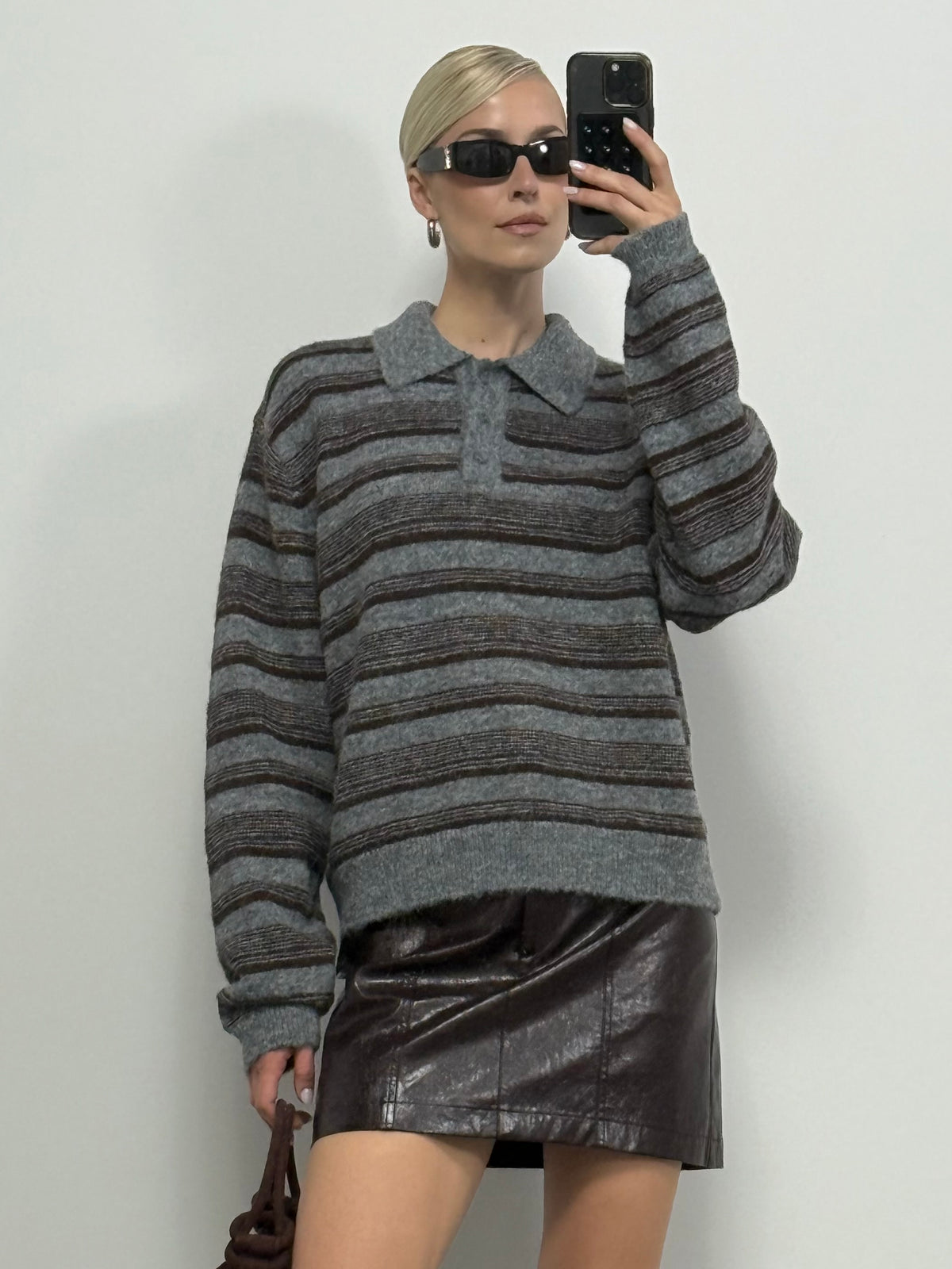 Eine Frau mit Sonnenbrille macht ein Selfie. Sie trägt den Pullover „Delia“ von LeGer by Lena Gercke mit einem eleganten Streifenmuster, dazu einen dunkelbraunen Lederminirock, und hält eine braune Tasche in der Hand, während sie vor einer schlichten, hellen Wand steht.