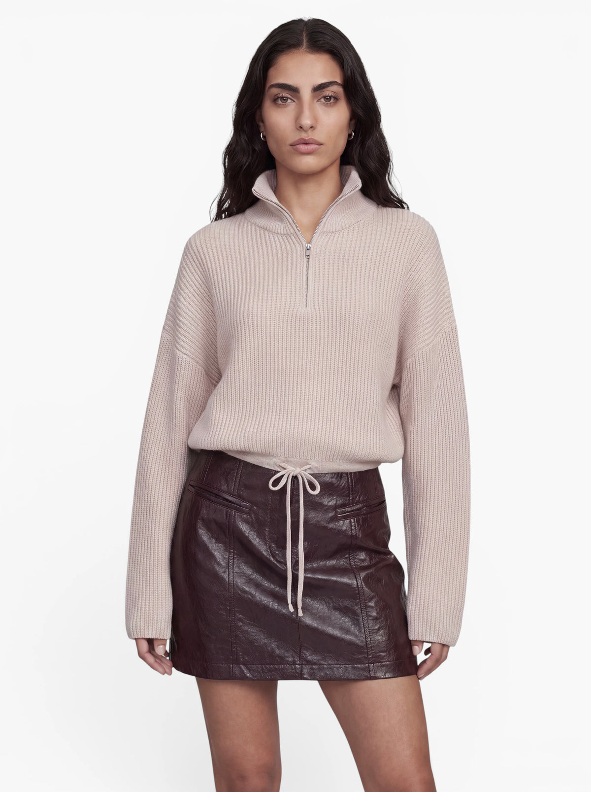 Eine Person steht vor einem weißen Hintergrund und trägt den Pullover „Michelle“ von LeGer by Lena Gercke – einen kurzen Strickpullover mit hohem Kragen und charakteristischer Rippenstruktur sowie einem Halbreißverschluss am Kragen –, kombiniert mit einem dunkelbraunen Lederminirock mit Kordelzug in der Taille.