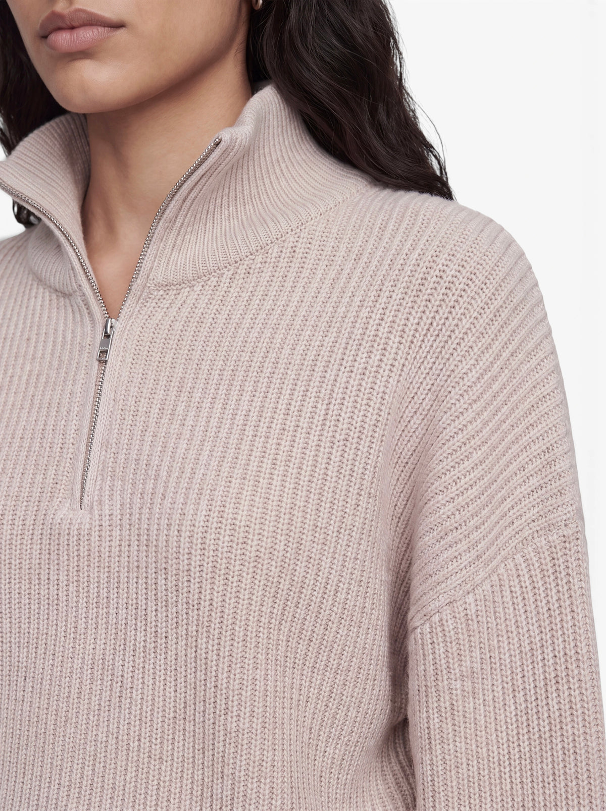Eine Frau ist von den Schultern aufwärts zu sehen. Sie trägt den Pullover „Michelle“ von LeGer by Lena Gercke – einen hellbeigen, kurzen Strickpullover mit Rippenstruktur, halbem Reißverschluss und hohem Kragen. Der Reißverschluss ist teilweise geöffnet und gibt den Blick auf ihr dunkles, welliges Haar frei.