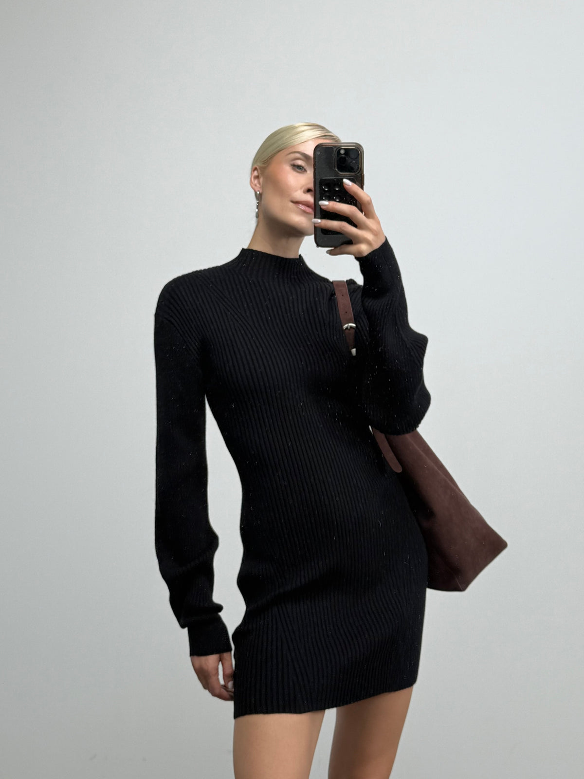 Eine Person mit blonden Haaren, die das langärmelige Strickkleid „Arvena“ mit Stehkragen von LeGer by Lena Gercke trägt und eine braune Tasche in der Hand hält, macht mit einem Smartphone vor einem einfarbigen, hellen Hintergrund ein Spiegel-Selfie.
