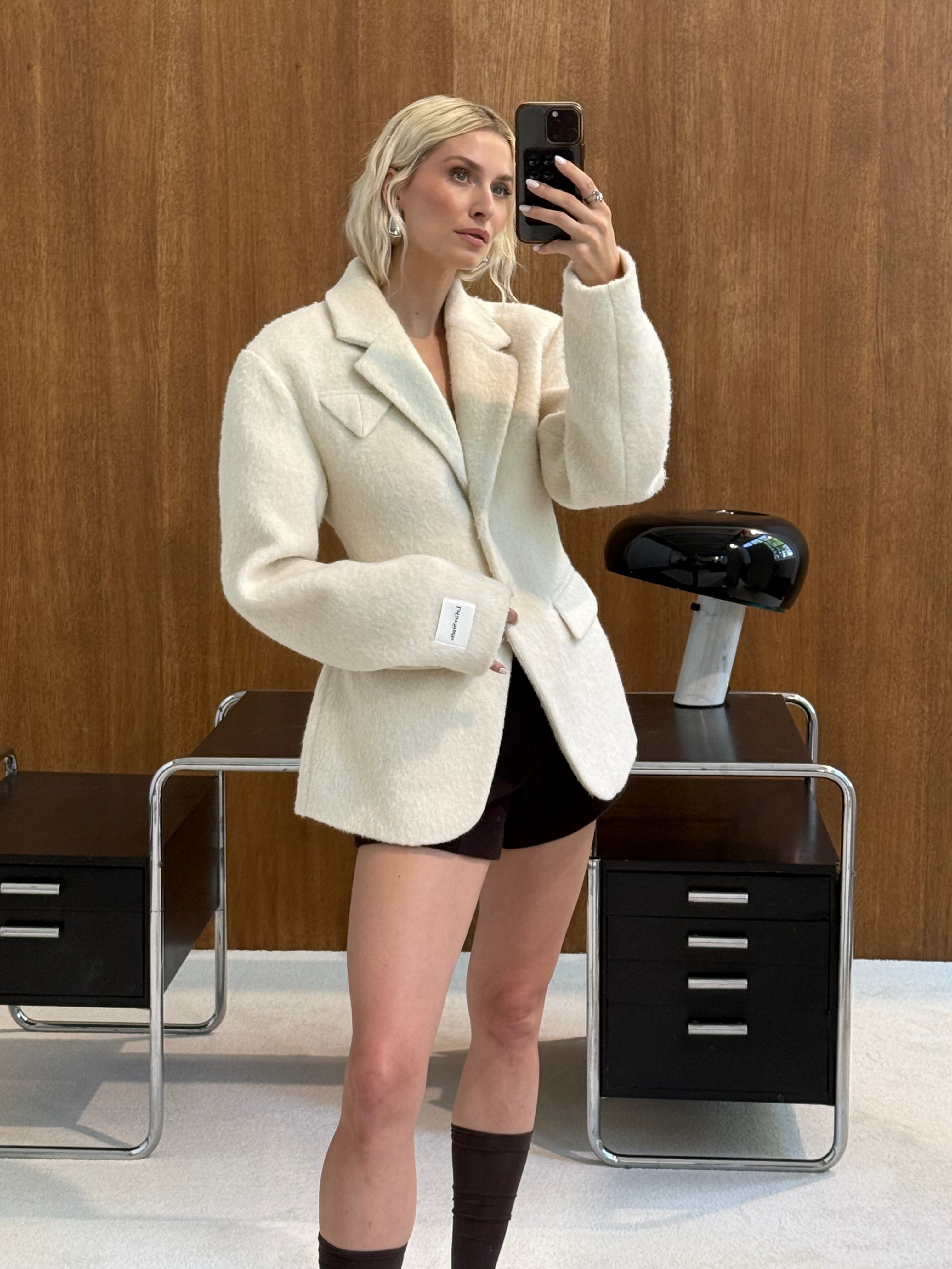 Eine Person, die die Jacke „Mila“ von LeGer by Lena Gercke in Cremefarbton sowie schwarze Shorts trägt, macht ein Spiegel-Selfie in einem modernen Büro mit Holzvertäfelung, schwarzen Schreibtischen und einer schwarzen Schreibtischlampe.