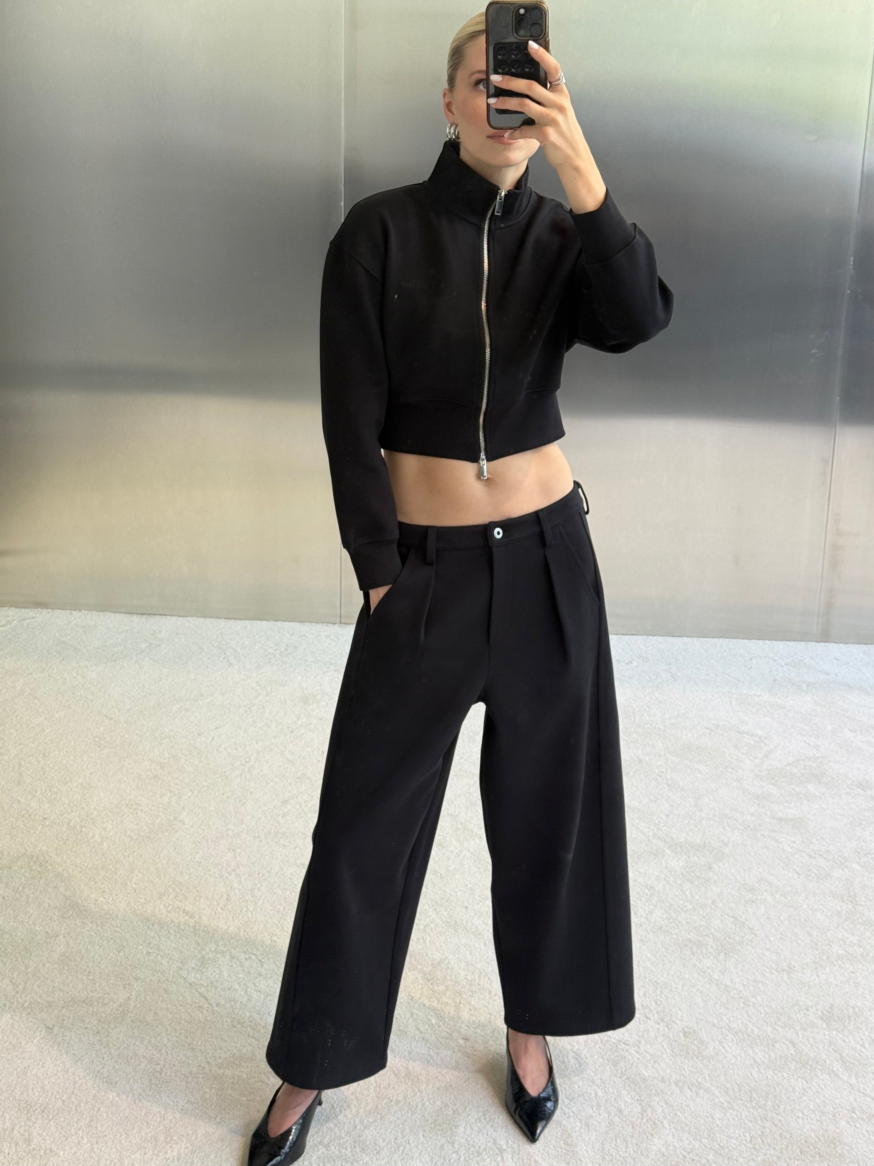 Eine Person steht drinnen auf einem hellen Teppich und macht mit ihrem Handy ein Spiegel-Selfie. Sie trägt eine kurze schwarze Jacke mit Reißverschluss, eine elegante Damenhose mit weitem Bein – genauer gesagt das Modell „Kasha“ von LeGer by Lena Gercke für besonders lange Beine – sowie schwarze Pumps.