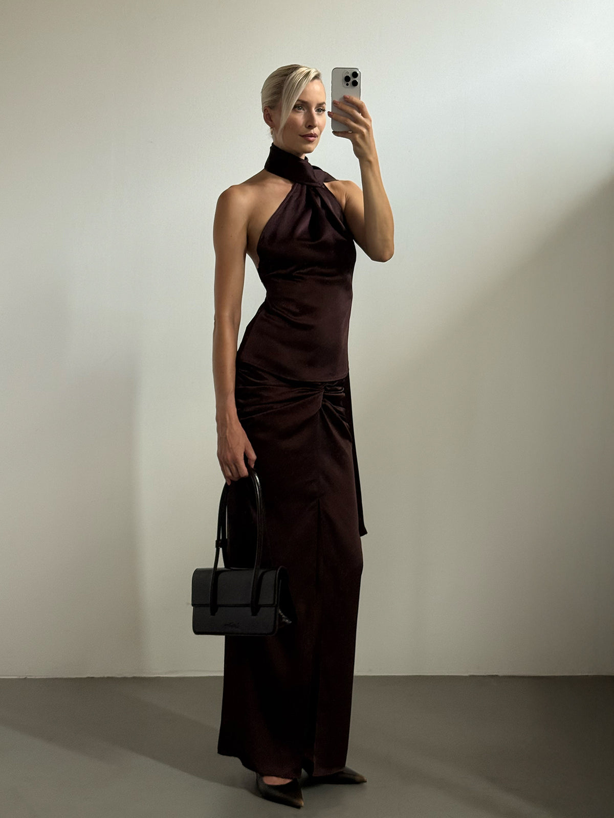Eine Frau trägt ein ärmelloses, dunkelbraunes Midi-Kleid der Marke Josephin mit hohem Ausschnitt und hält eine kleine schwarze Handtasche in der Hand.