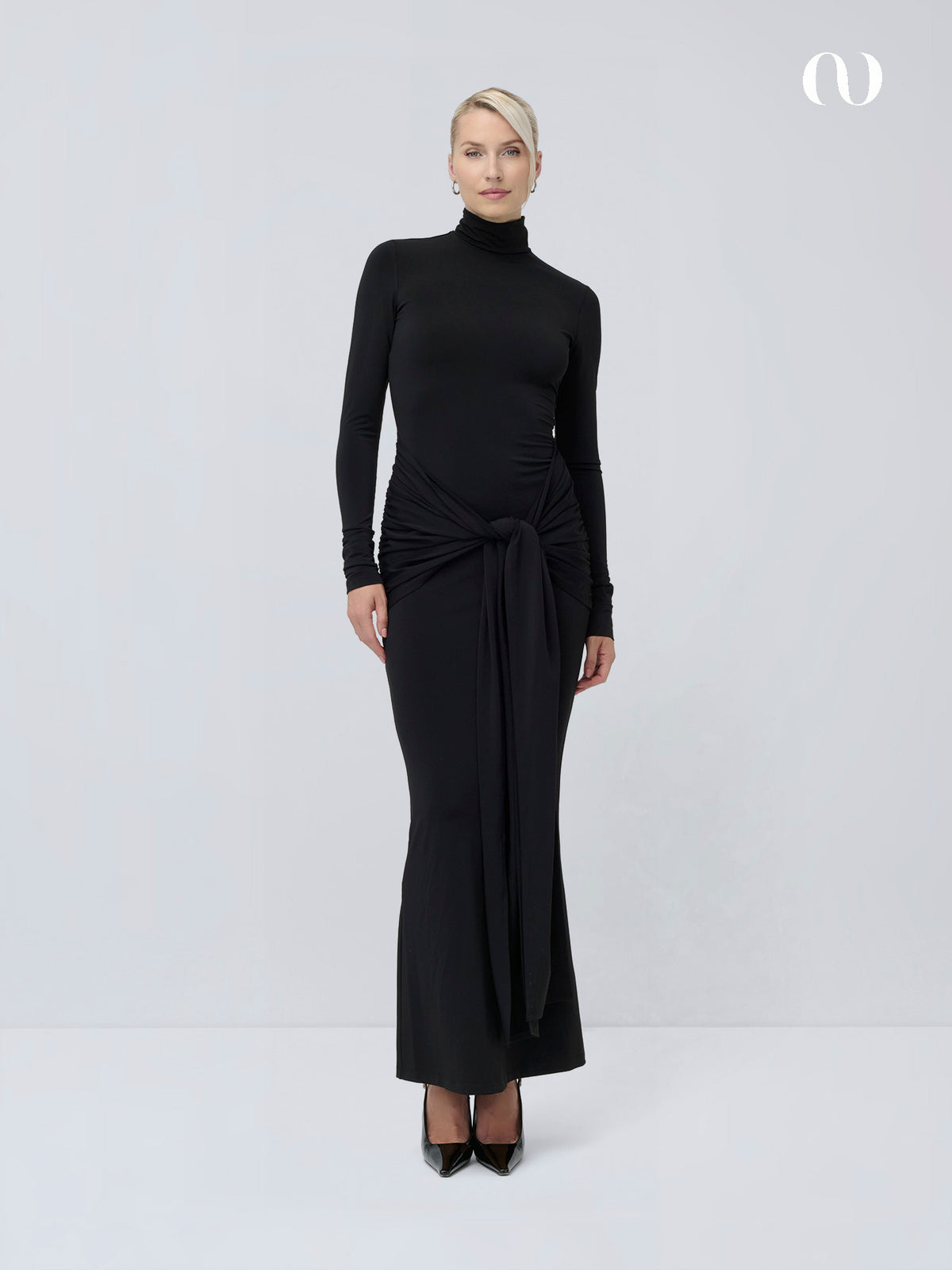 Eine Frau, die das schwarze „Izzie“-Kleid trägt – ein langärmeliges Maxikleid mit einem drapierten Knoten an der Taille –, dazu schwarze High Heels.