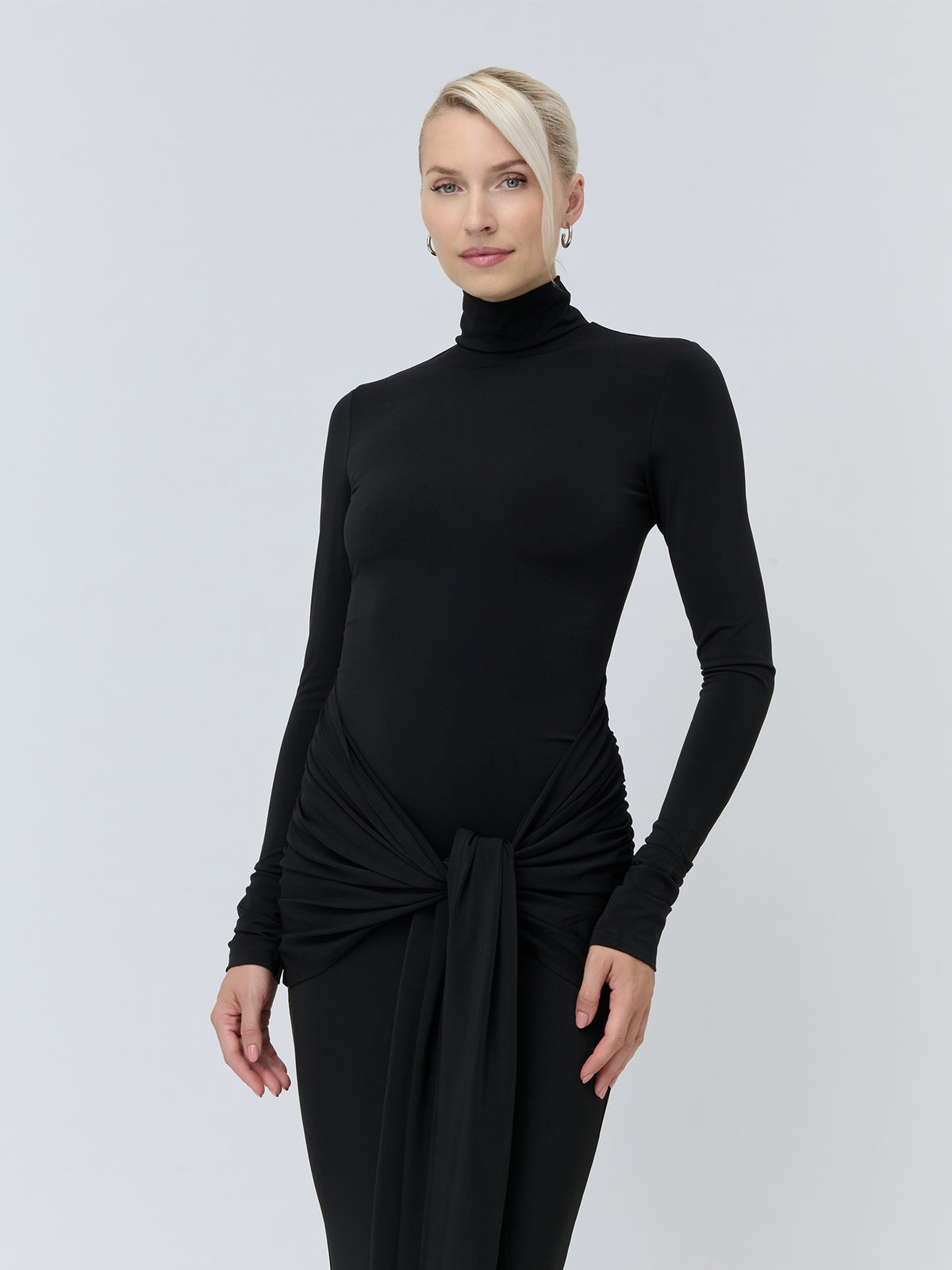 Eine Person, die ein schwarzes, langärmeliges „Izzie“-Kleid mit Rollkragen, bodenlangem Saum und einem geknoteten Bindeband an der Taille trägt.