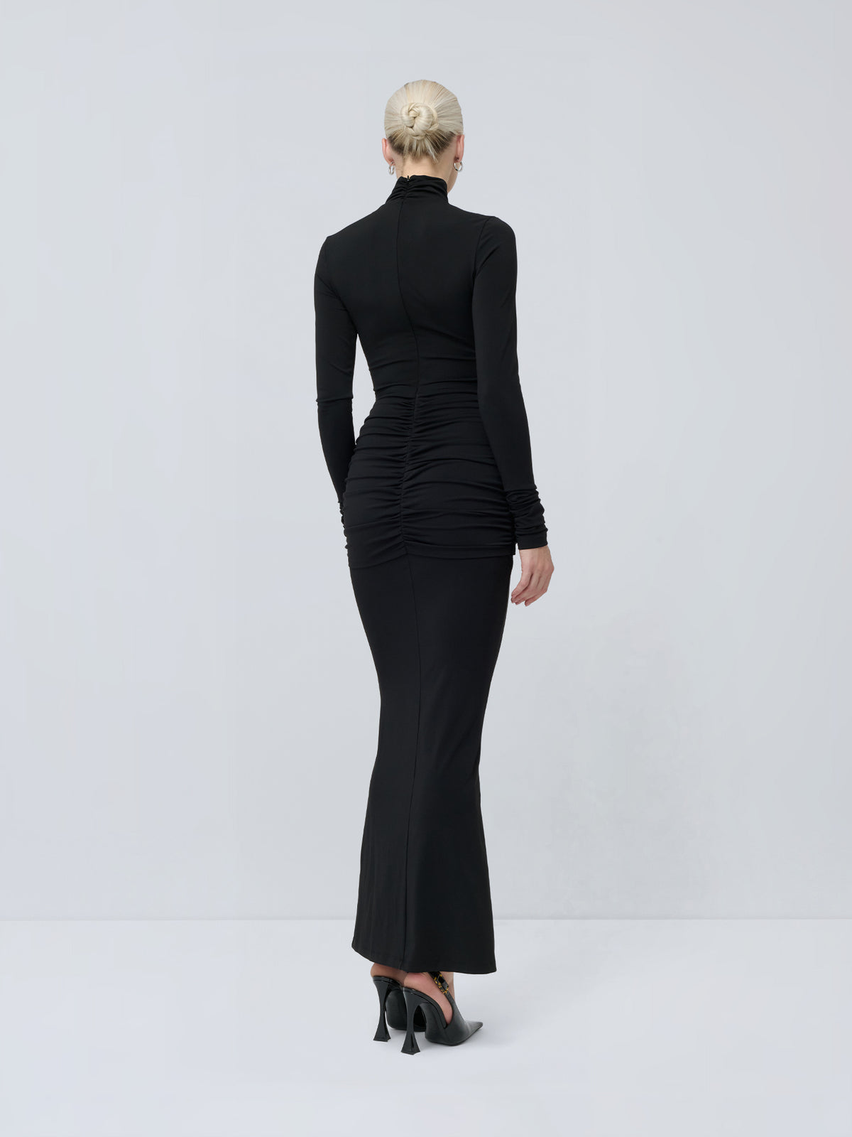 Rückansicht einer Person, die ein langärmeliges, schwarzes „Izzie“-Kleid von LeGer by Lena Gercke mit Stehkragen und eleganter Drapierung trägt, kombiniert mit schwarzen Mules mit hohen Absätzen.