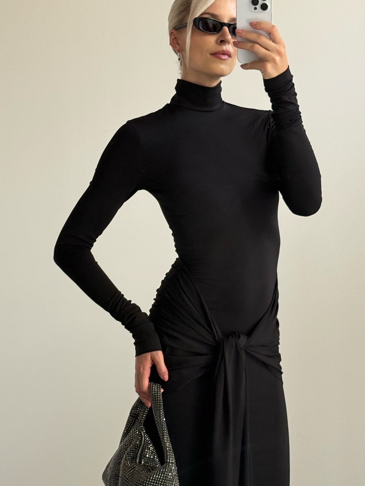 Eine Person, die ein schwarzes, langärmeliges „Izzie“-Kleid von LeGer by Lena Gercke mit geknoteter Drapierung an der Taille und eine schwarze Sonnenbrille trägt, macht ein Spiegel-Selfie, während sie eine glitzernde Handtasche in der Hand hält.
