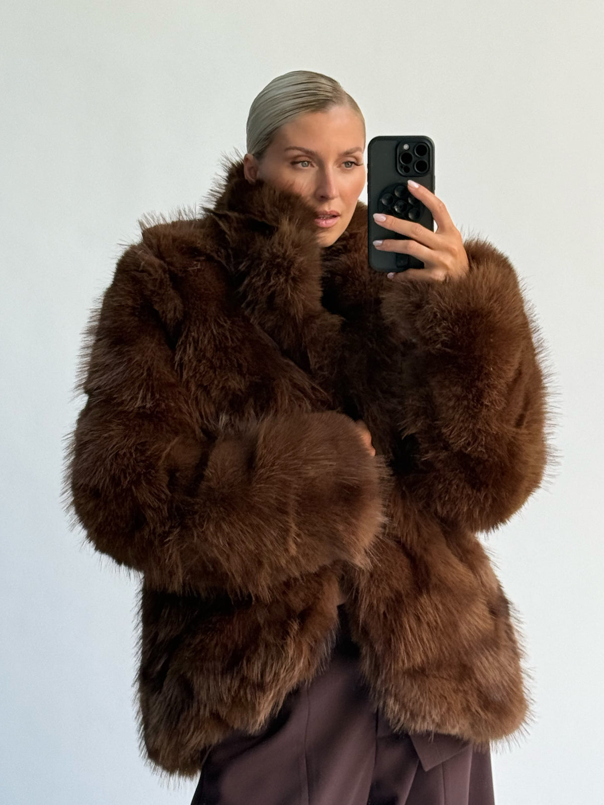 Eine Person, die ein Spiegel-Selfie macht und dabei eine dunkelbraune „Dion“-Jacke aus Kunstfell von LeGer by Lena Gercke mit offener Vorderseite trägt.