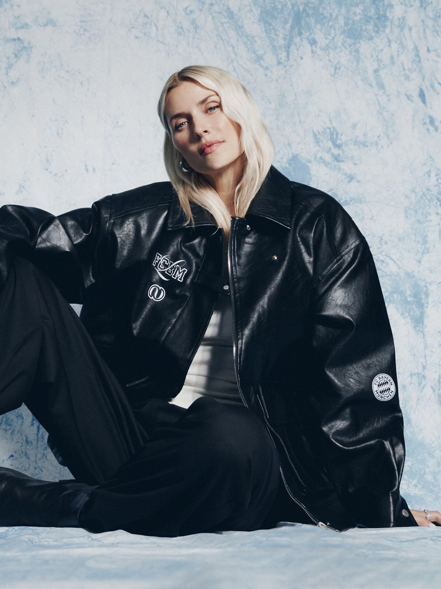 Eine Person mit langen blonden Haaren sitzt vor einem hellblau strukturierten Hintergrund, trägt die LeGer by Lena Gercke Elisa Jacket, eine schwarze Hose und ein weißes Oberteil und blickt selbstbewusst in die Kamera.