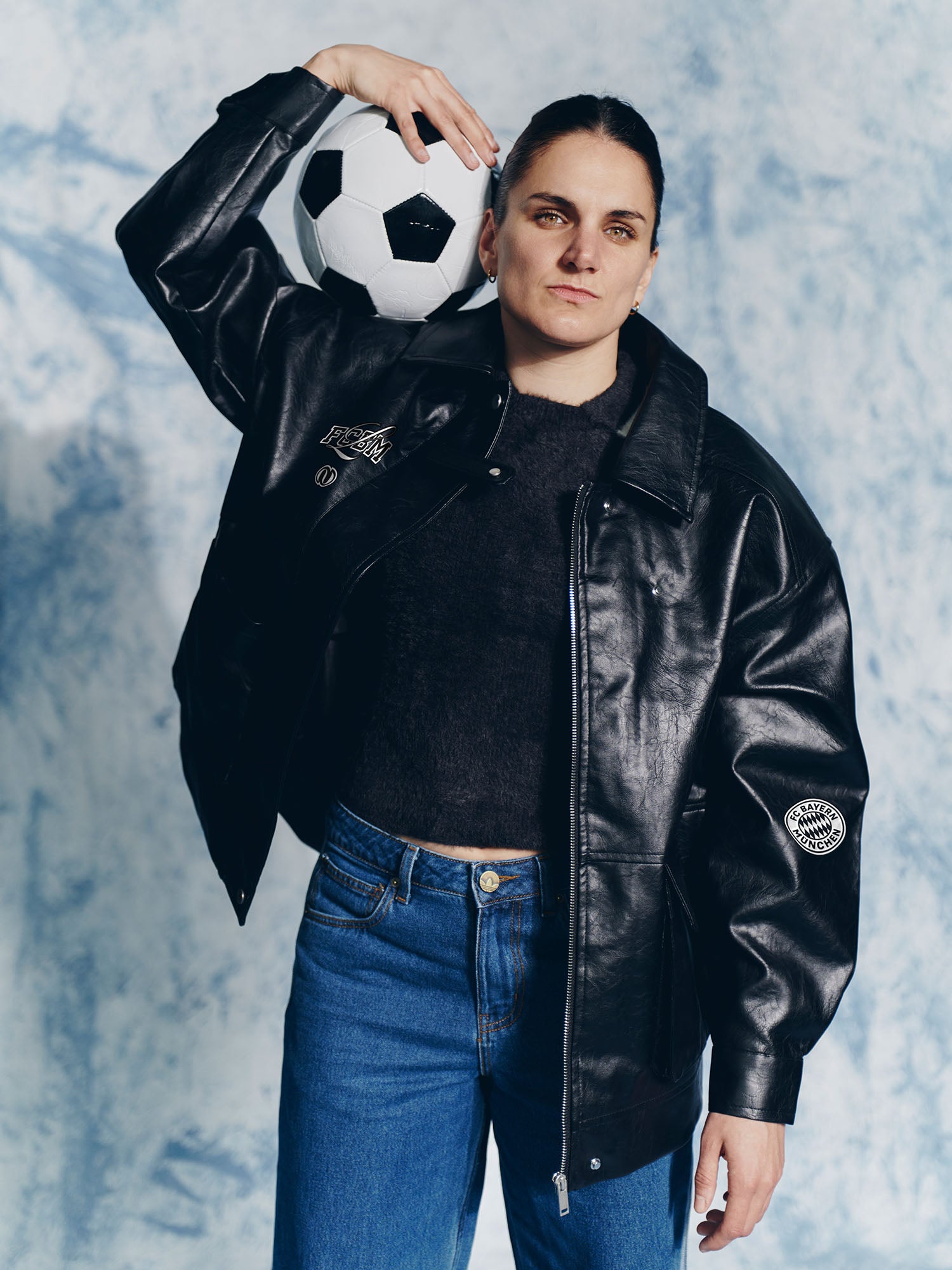 Eine Person trägt eine LeGer by Lena Gercke Elisa Jacke und eine blaue Jeans und hält einen Fußball auf ihrer Schulter vor einem hellblau-weiß strukturierten Hintergrund, der an den Stil der FC Bayern Frauen erinnert.