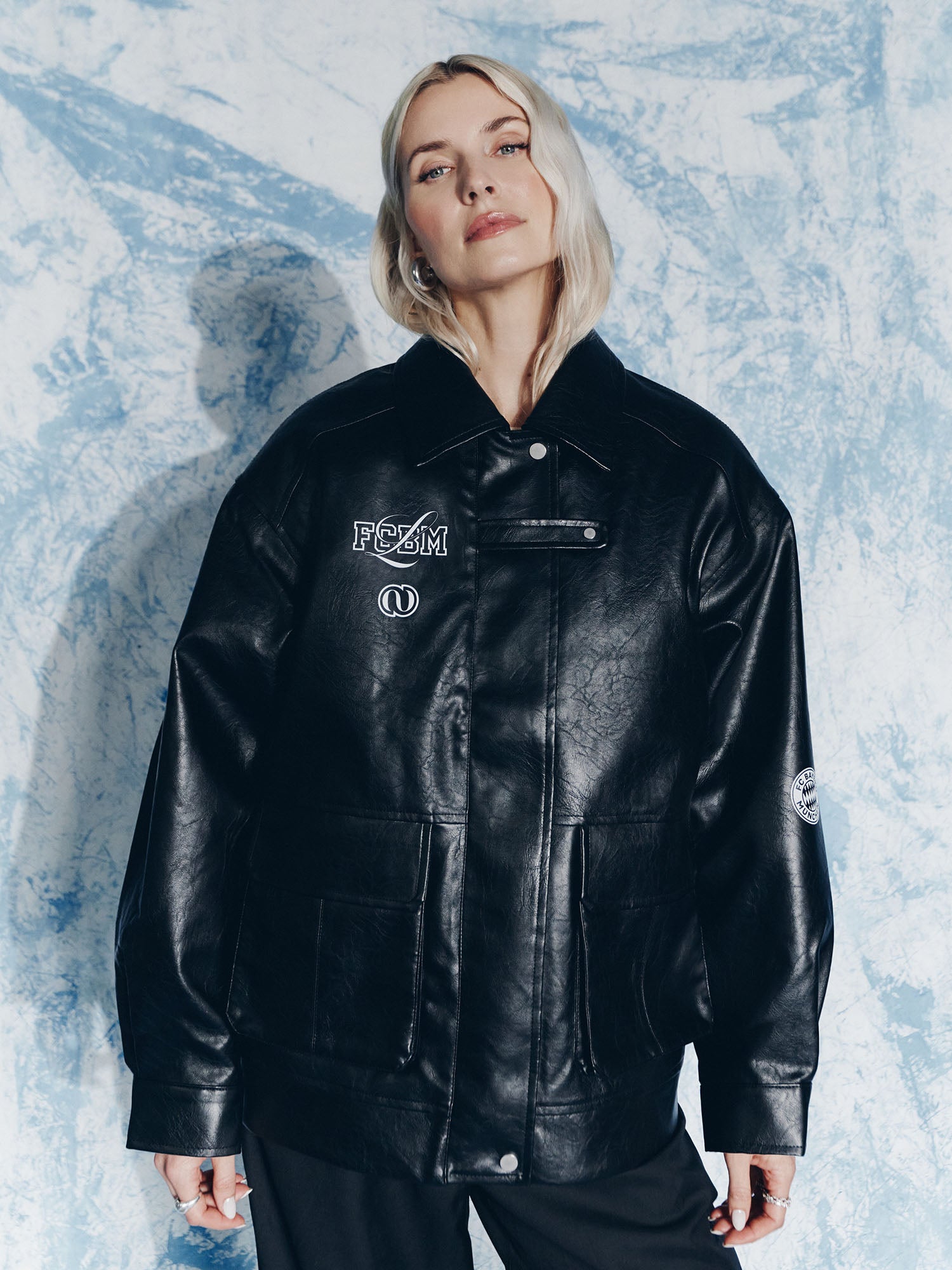 Eine Frau mit hellblondem Haar trägt eine locker sitzende schwarze Elisa Jacket von LeGer by Lena Gercke mit weißen Logos und Aufnähern vor einem strukturierten blau-weißen Hintergrund.