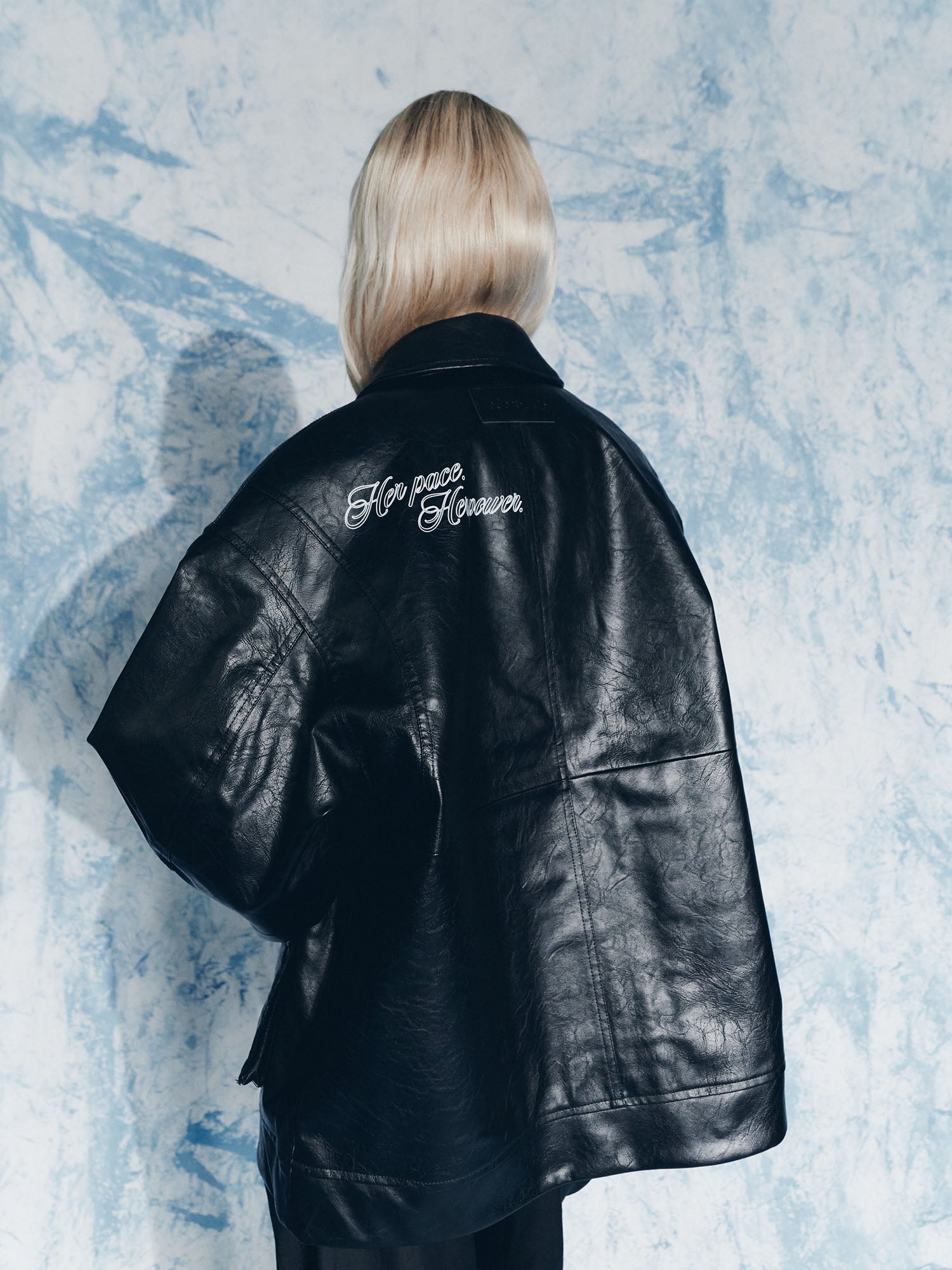 Eine Person mit langem, hellblondem Haar blickt in die Ferne. Sie trägt die übergroße Jacke Elisa von LeGer by Lena Gercke mit der Stickerei "For rose Flower" auf dem Rücken vor einem blau-weiß marmorierten Hintergrund.