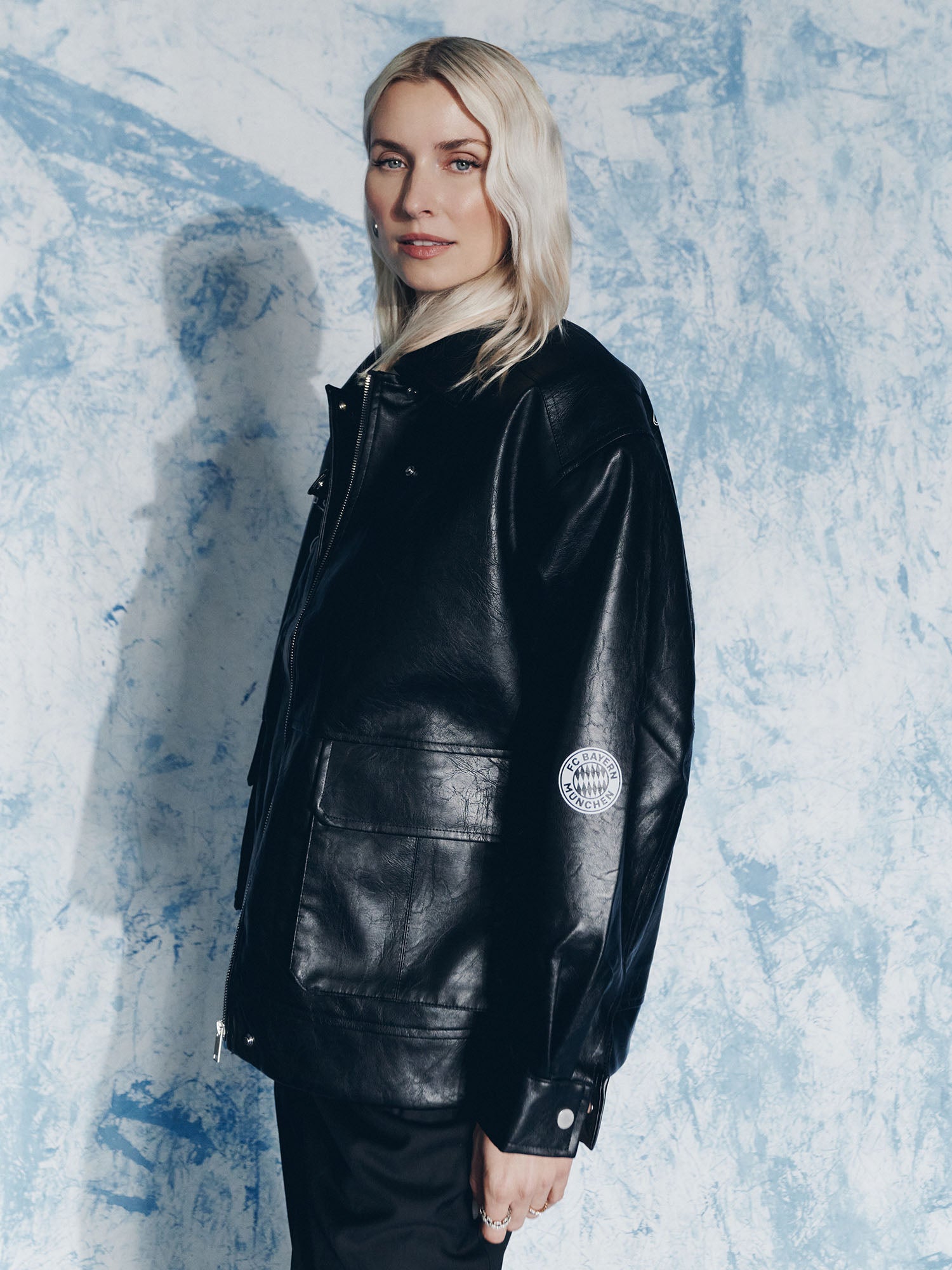 Eine Person mit langem, hellblondem Haar trägt die Jacke Elisa von LeGer by Lena Gercke, steht vor einem blau-weiß strukturierten Hintergrund und blickt mit einem leichten Lächeln in die Kamera.