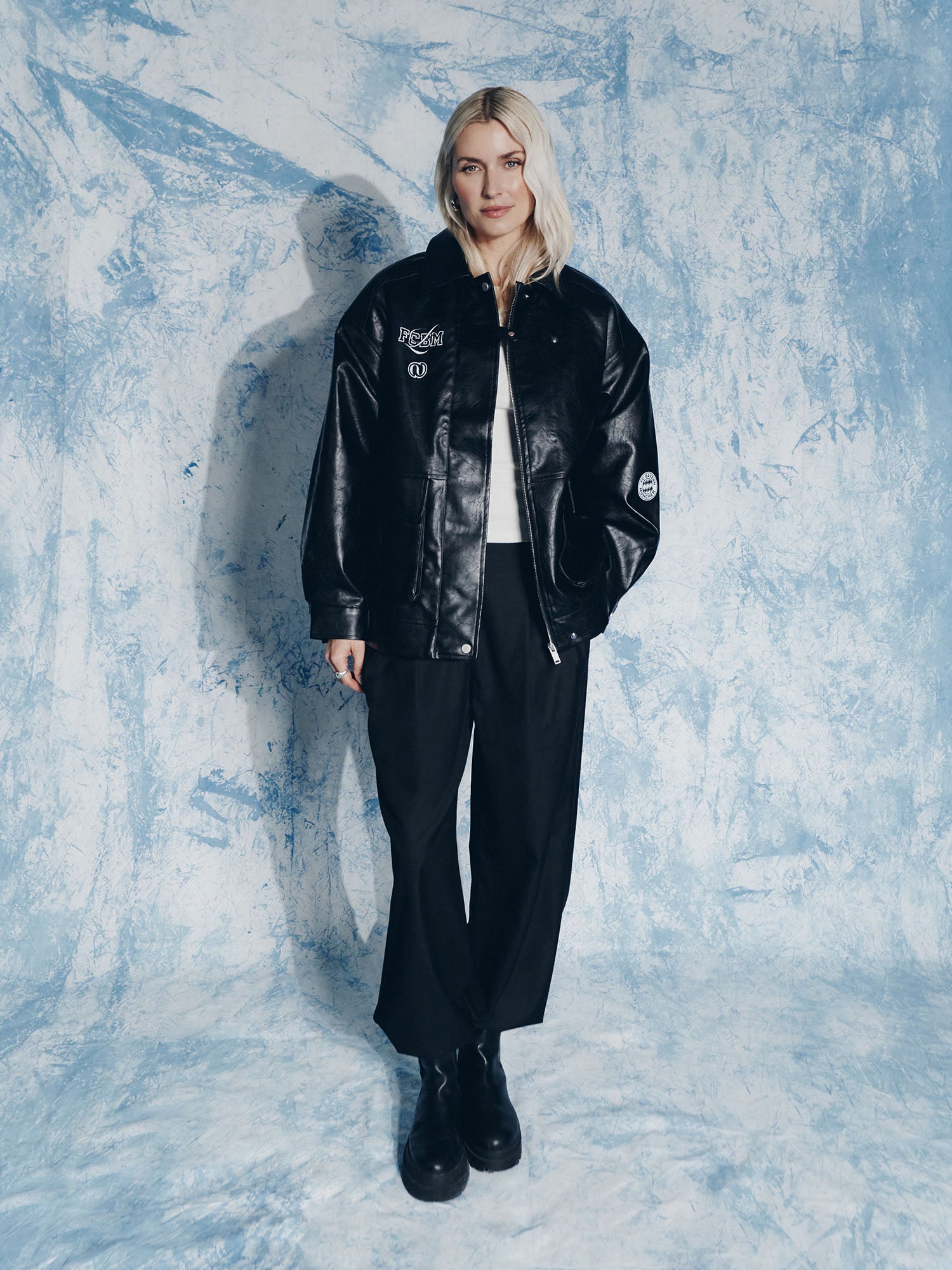 Eine blonde Frau steht vor einem blau-weiß strukturierten Hintergrund und trägt die LeGer by Lena Gercke Elisa Jacket, eine schwarze Hose mit weitem Bein, schwarze Plateaustiefel und ein weißes Oberteil. Sie blickt mit neutralem Blick in die Kamera.