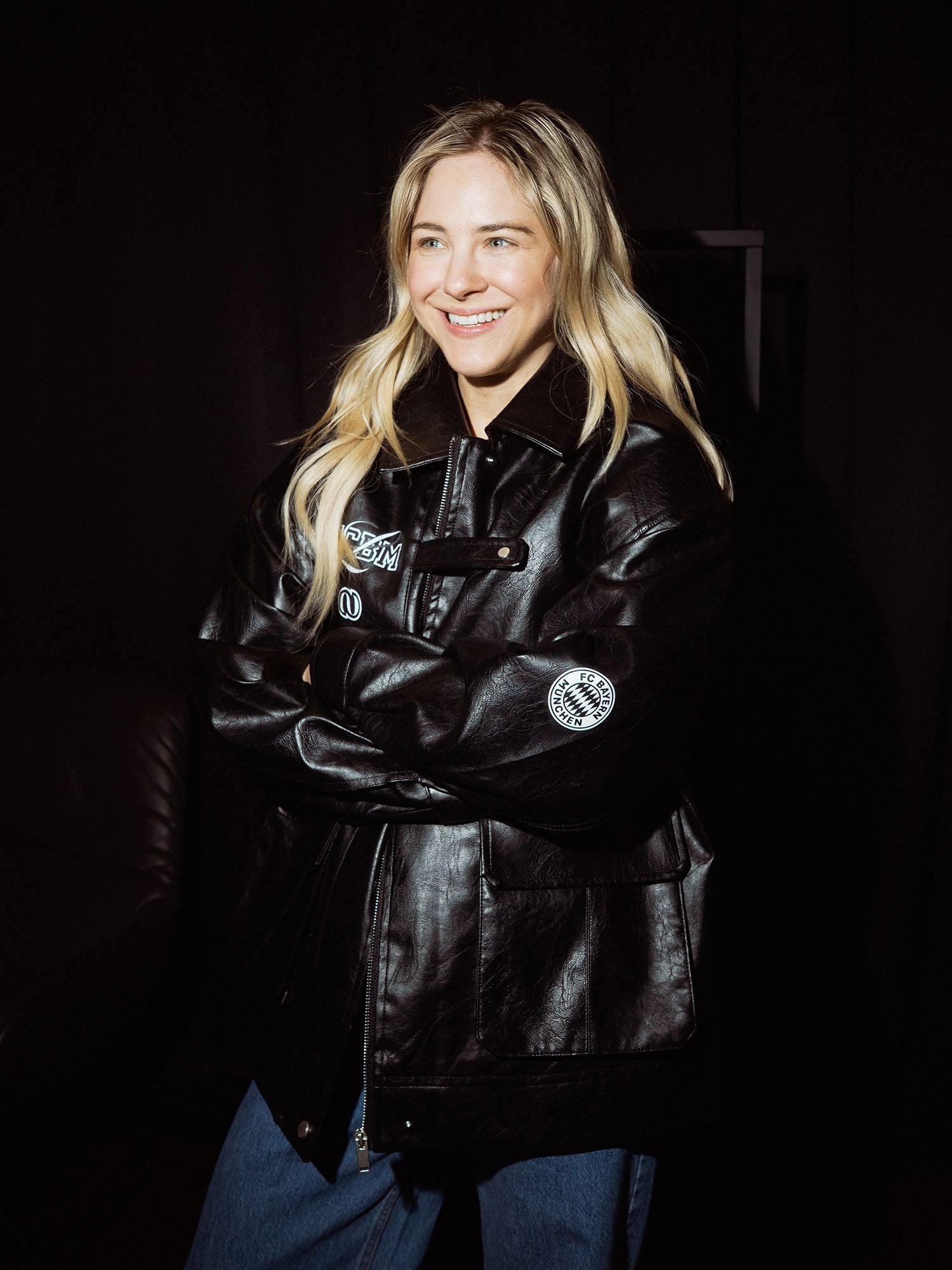 Eine Frau mit langen blonden Haaren, die mit verschränkten Armen lächelt, trägt eine schwarze Elisa Jacket von LeGer by Lena Gercke und blaue Jeans vor einem dunklen Hintergrund.