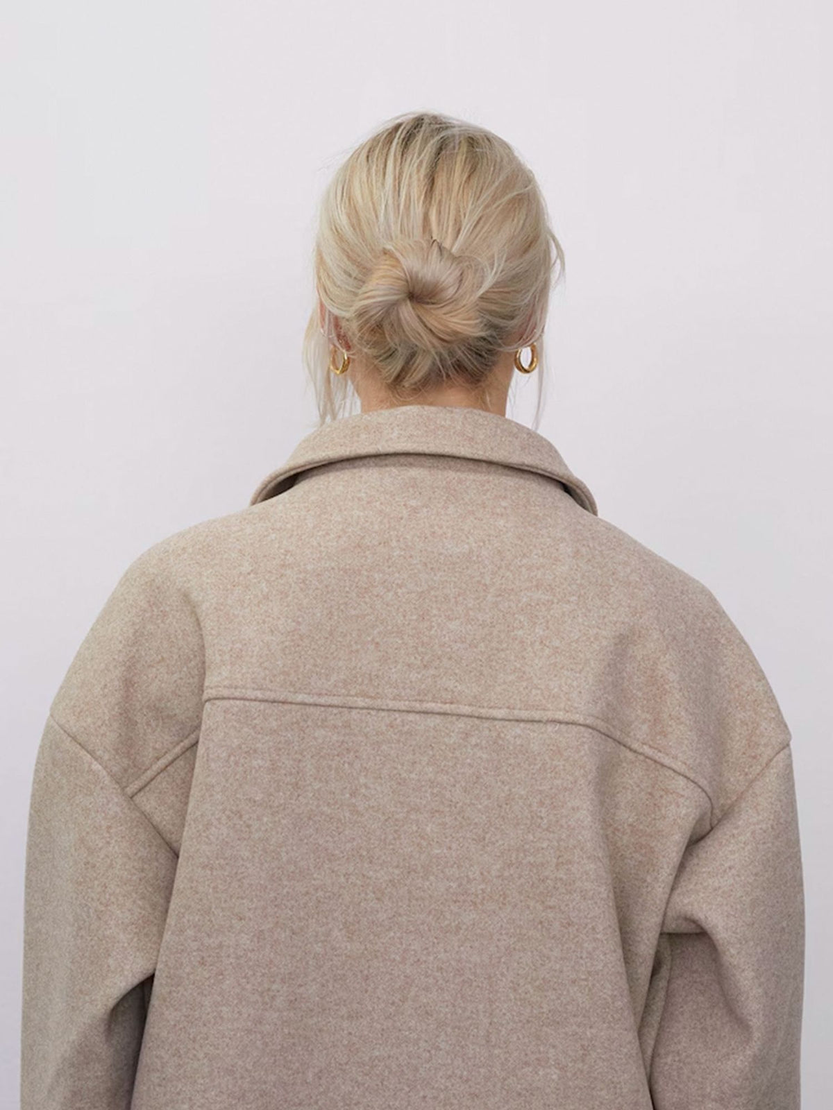 Eine Person mit blonden Haaren, die zu einem tief sitzenden Dutt zusammengebunden sind, trägt goldene Creolen und die beige „Eva“-Jacke mit Brusttaschen von LeGer by Lena Gercke; sie ist von hinten vor einem schlichten, hellen Hintergrund zu sehen.