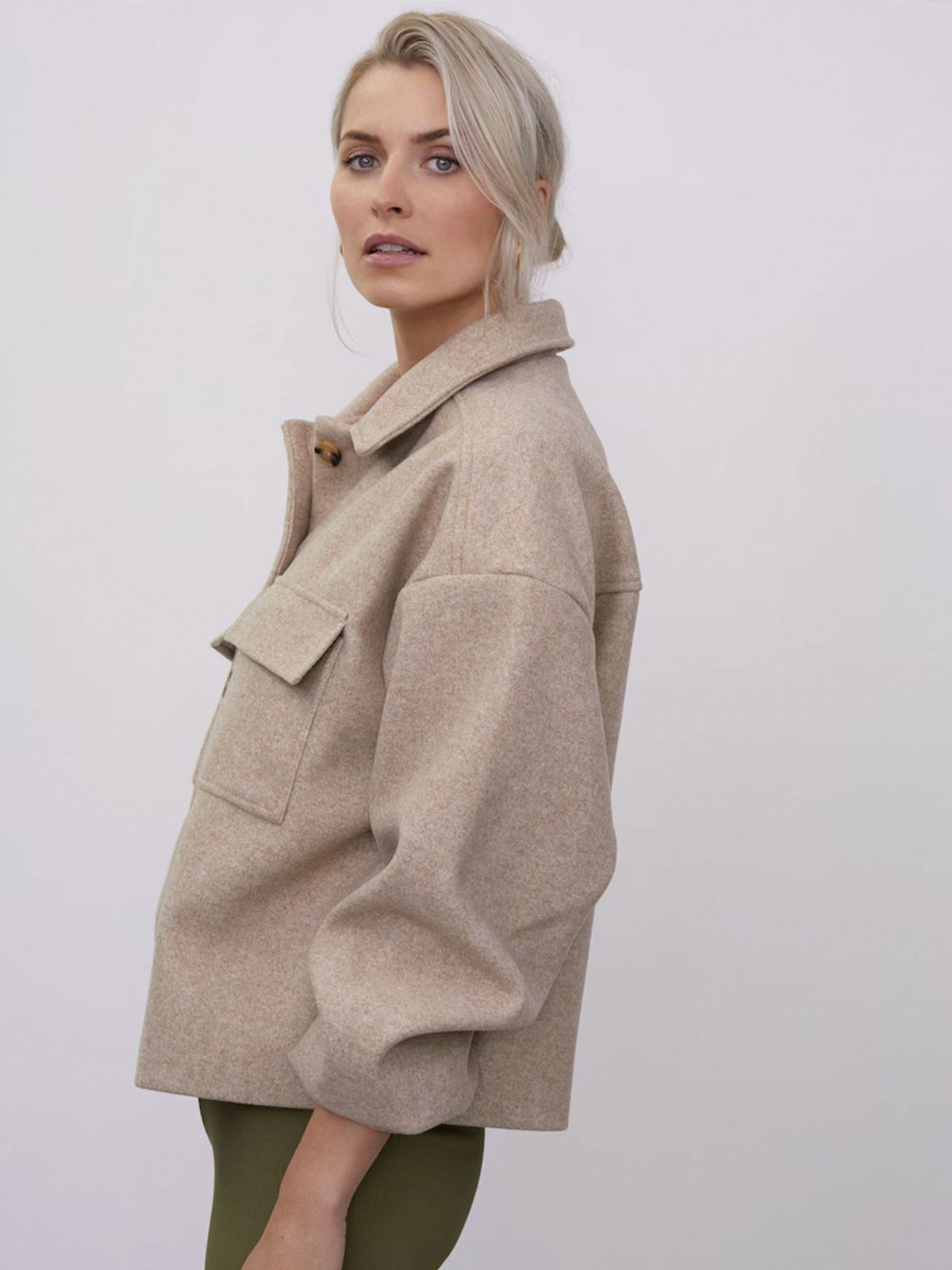 Eine Frau mit hellblondem Haar, die die beige „Eva“-Jacke von LeGer by Lena Gercke mit großen Brusttaschen trägt, steht seitlich und blickt vor einem schlichten, hellen Hintergrund in die Kamera.