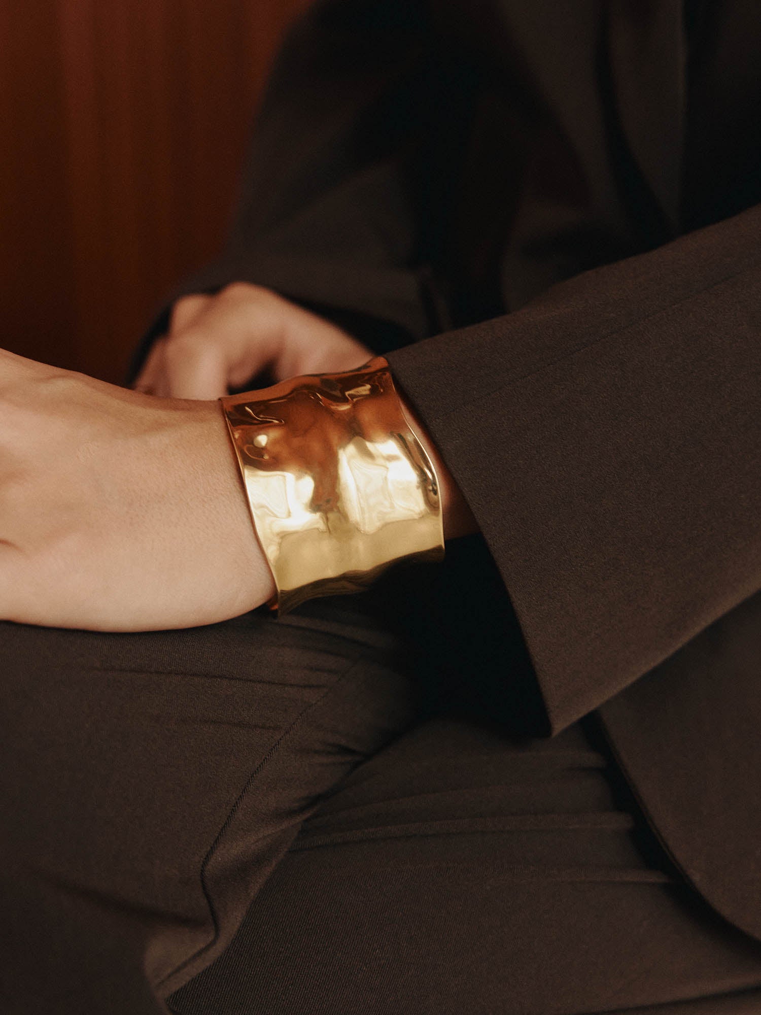 Eine Person in einem dunklen Anzug stützt ihre Hand auf ihr Knie und hebt die LeGer x Purelei - Modern Muse Armfessel von LeGer by Lena Gercke hervor - ein kühnes, goldglänzendes Statement-Stück.